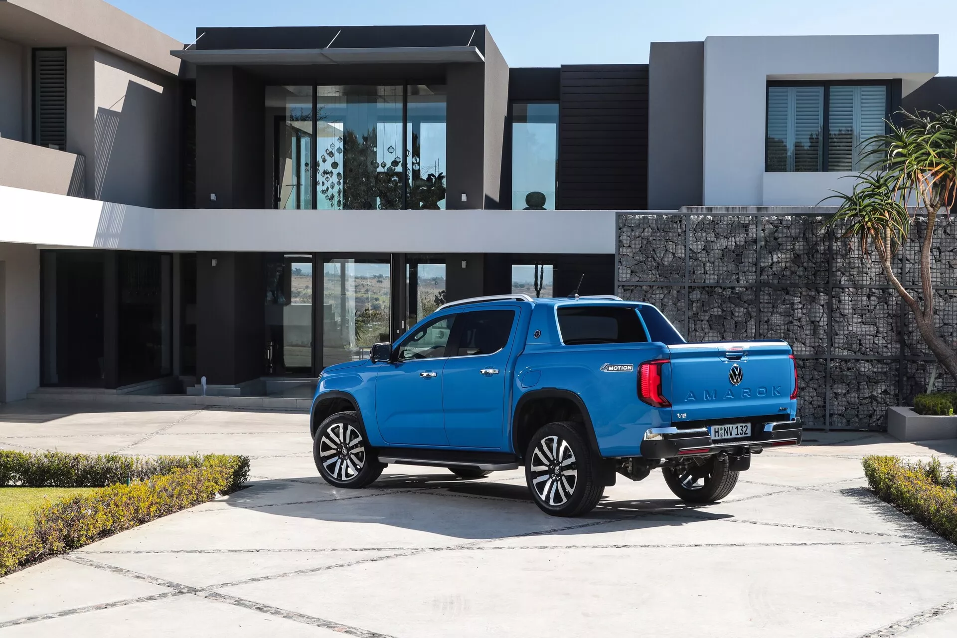 Vue arrière trois-quarts d'un pick-up bleu moderne VW Nutzfahrzeuge Amarok 2023, présentant ses roues en alliage, sa peinture métallisée brillante, ses capteurs arrière, ses feux arrière à LED et son antenne aileron de requin. Le véhicule est garé sur une allée pavée devant une résidence contemporaine luxueuse, soulignant son utilité dans un environnement résidentiel haut de gamme.