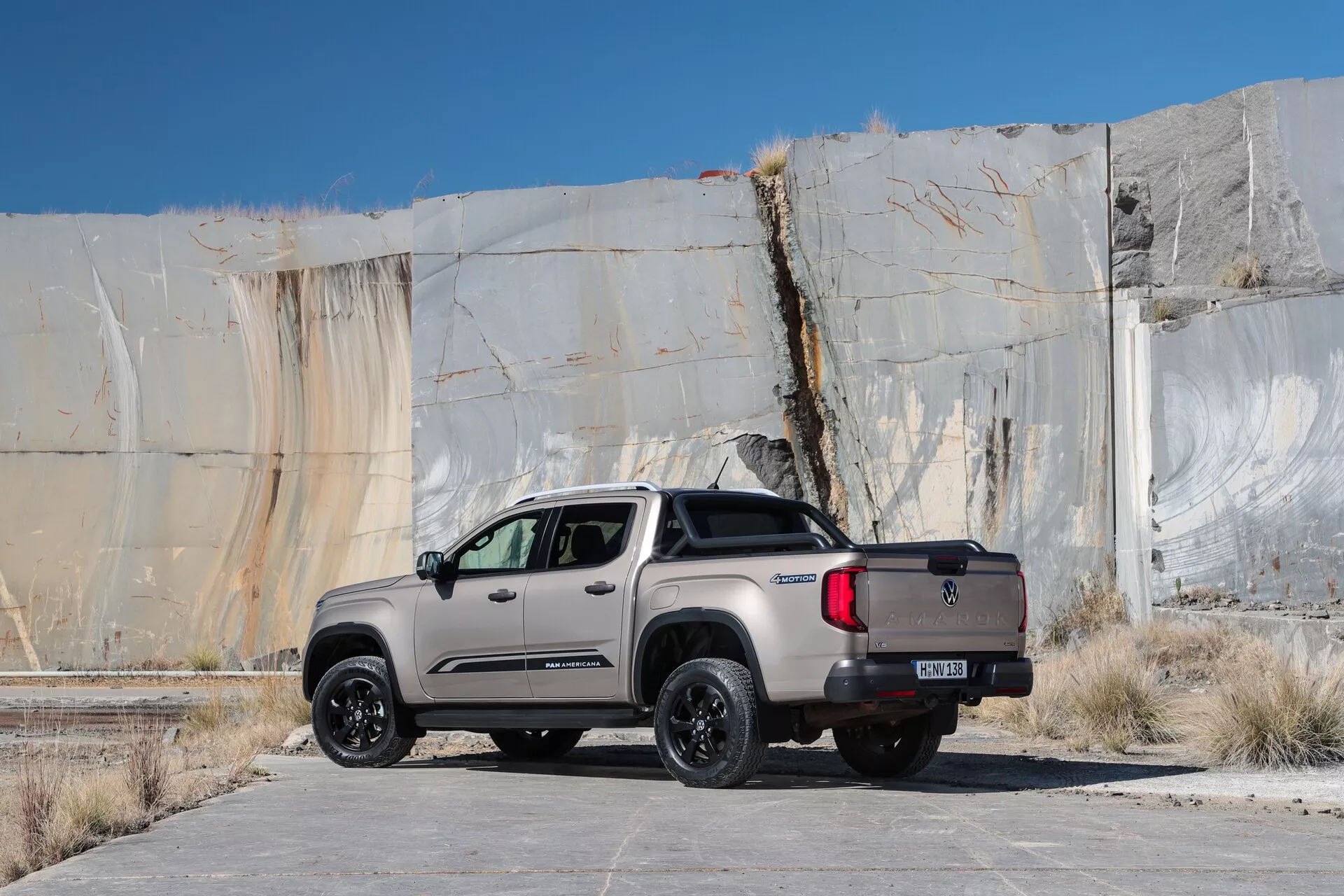 Vue arrière trois-quarts d'un pick-up VW Nutzfahrzeuge Amarok 2023 (2ème génération), montrant ses éléments de design latéraux et arrière. Le camion est garé sur une surface de béton plate contre un décor rugueux et texturé. Il affiche une couleur de peinture mate grise ou beige avec des accents noirs sur les roues, rétroviseurs et garnitures, suggérant une édition spéciale ou un package tout-terrain. Les caractéristiques incluent des roues en alliage, des pneus tout-terrain, une configuration à double cabine avec quatre portes et un hayon arrière avec badge du fabricant et attelage de remorque. Les feux arrière LED et le couvre-benne contribuent à son design moderne et utilitaire.