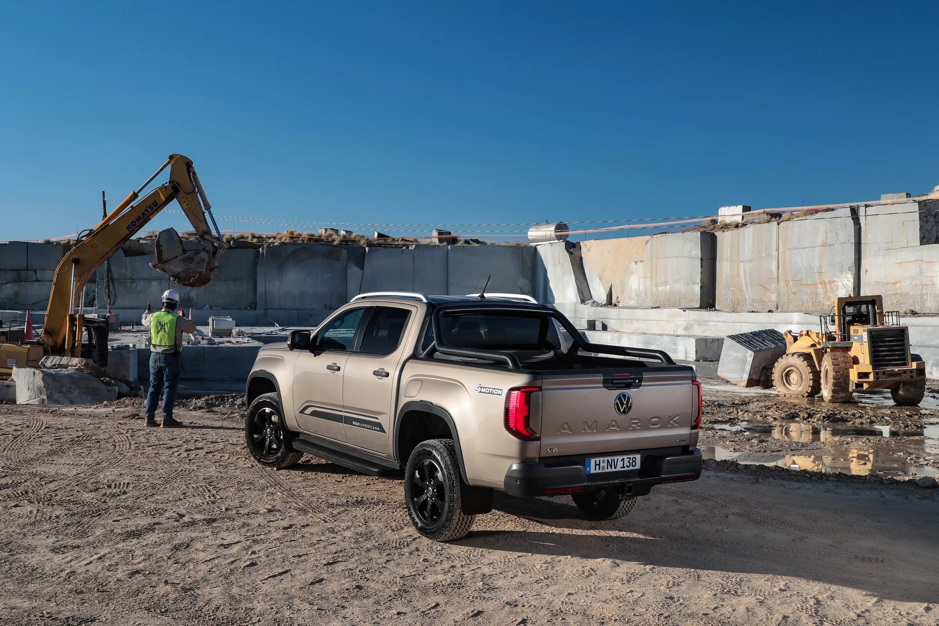 Image d'un pick-up VW Nutzfahrzeuge Amarok 2023 dans un environnement industriel avec des équipements d'excavation et de grands blocs de pierre en arrière-plan. Capturé sous un angle trois-quarts arrière, le camion de couleur claire avec des garnitures et des roues noires est montré en entier avec une double cabine et un plateau couvert. Une personne en gilet haute visibilité se trouve à l'arrière-plan, renforçant le cadre de chantier de construction.