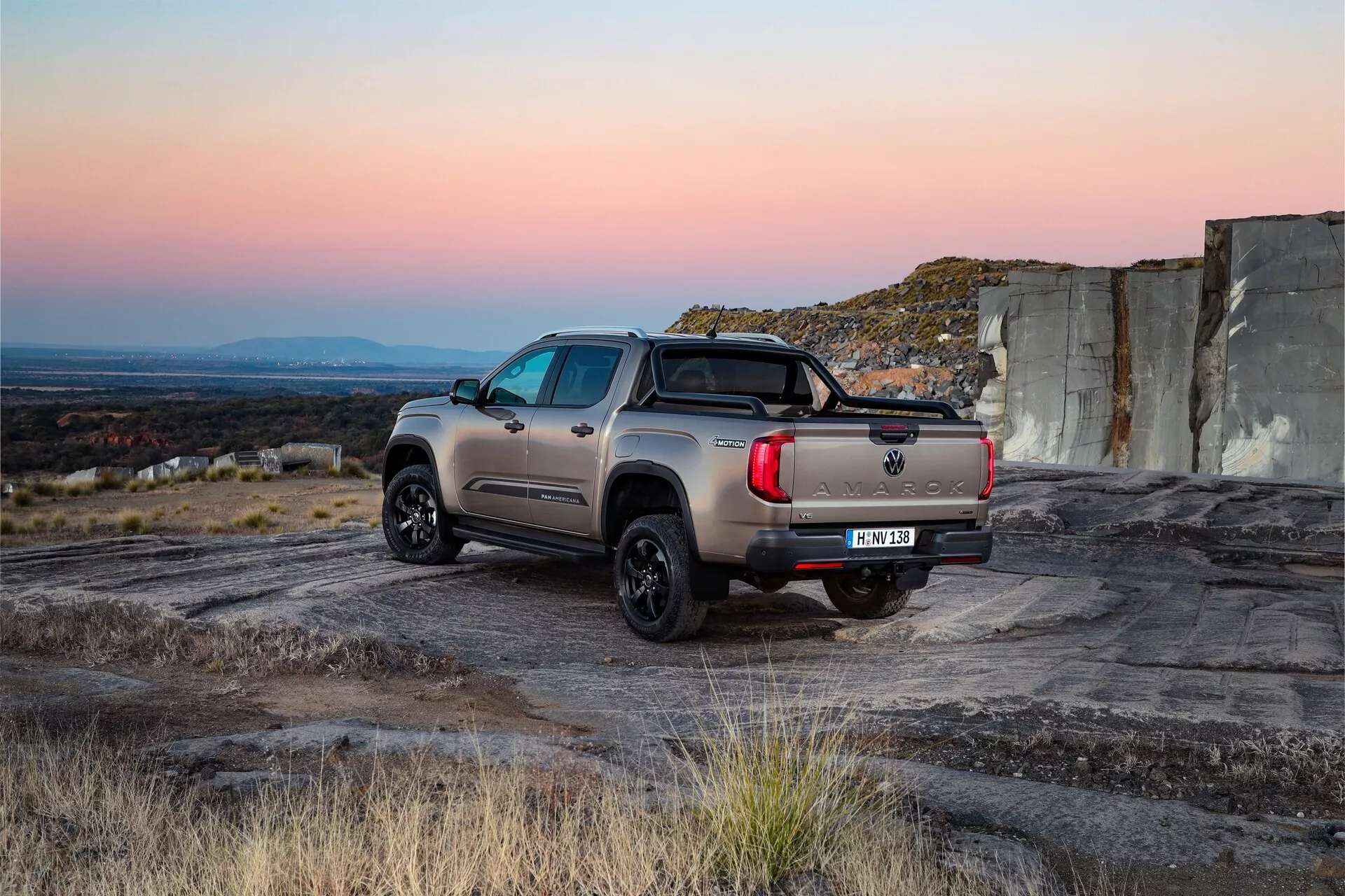 Vue arrière trois quarts du VW Nutzfahrzeuge Amarok 2023, mettant en valeur son design extérieur robuste sur un terrain accidenté. Le pickup moderne présente une carrosserie robuste avec une peinture non métallique, des feux arrière LED contemporains et un attelage de remorque sous le pare-chocs arrière. Les roues en alliage peintes en noir équipées de pneus tout-terrain soulignent son utilité robuste, tandis que la barre de sport dans la benne du camion ajoute un attrait esthétique et une fonctionnalité pour sécuriser le chargement. Les marchepieds latéraux et les vitres arrière teintées améliorent l'accessibilité et l'intimité. La position du véhicule dans une lumière naturelle aux teintes chaudes et aux ombres froides met en valeur ses attributs de performance et d'utilité.
