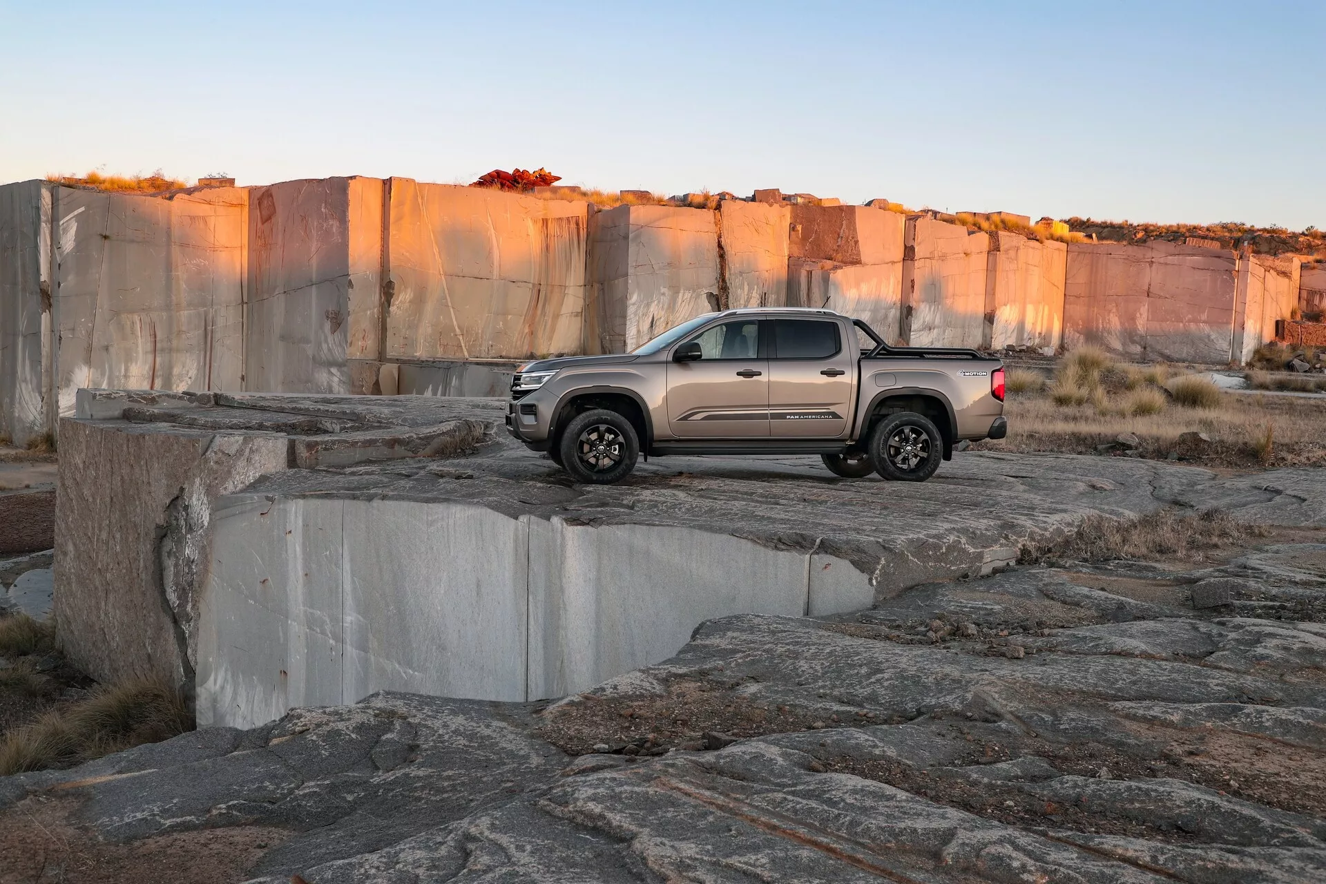 Pick-up de taille moyenne VW Nutzfahrzeuge Amarok 2023 (2ème génération) sur un terrain rocheux, probablement une mine à ciel ouvert. Le véhicule est photographié sous un angle arrière trois quarts à la fin de l'après-midi. L'image met en avant la configuration de cabine double, une couleur claire et atténuée, un design moderne avec des feux arrière LED, un hayon sculpté, des roues distinctives et des pneus à bande de roulement agressive, soulignant ses capacités tout-terrain.