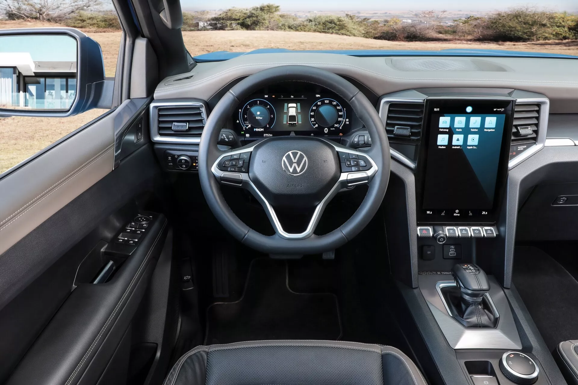 L'intérieur du Volkswagen Nutzfahrzeuge Amarok 2023 (2e génération) capturé depuis la perspective du conducteur, mettant en avant le tableau de bord, le volant et la console centrale. Le volant à trois branches comprend des commandes intégrées pour l'audio, le régulateur de vitesse et d'autres systèmes, tandis que le tableau de bord numérique affiche les informations de conduite essentielles. La console centrale inclut un système d'infodivertissement à écran tactile en orientation portrait pour la navigation et les paramètres du véhicule, entouré de boutons et de commandes physiques pour le contrôle du climat. En dessous, les commandes de la boîte de vitesses et des modes de conduite sont visibles, ainsi qu'un petit espace de rangement. Le design irradie de modernité avec des matériaux en cuir ou doux au toucher et des accents métalliques. La porte du conducteur montre les commandes des fenêtres et des rétroviseurs, avec un aperçu cohérent de la sellerie sur le siège passager.