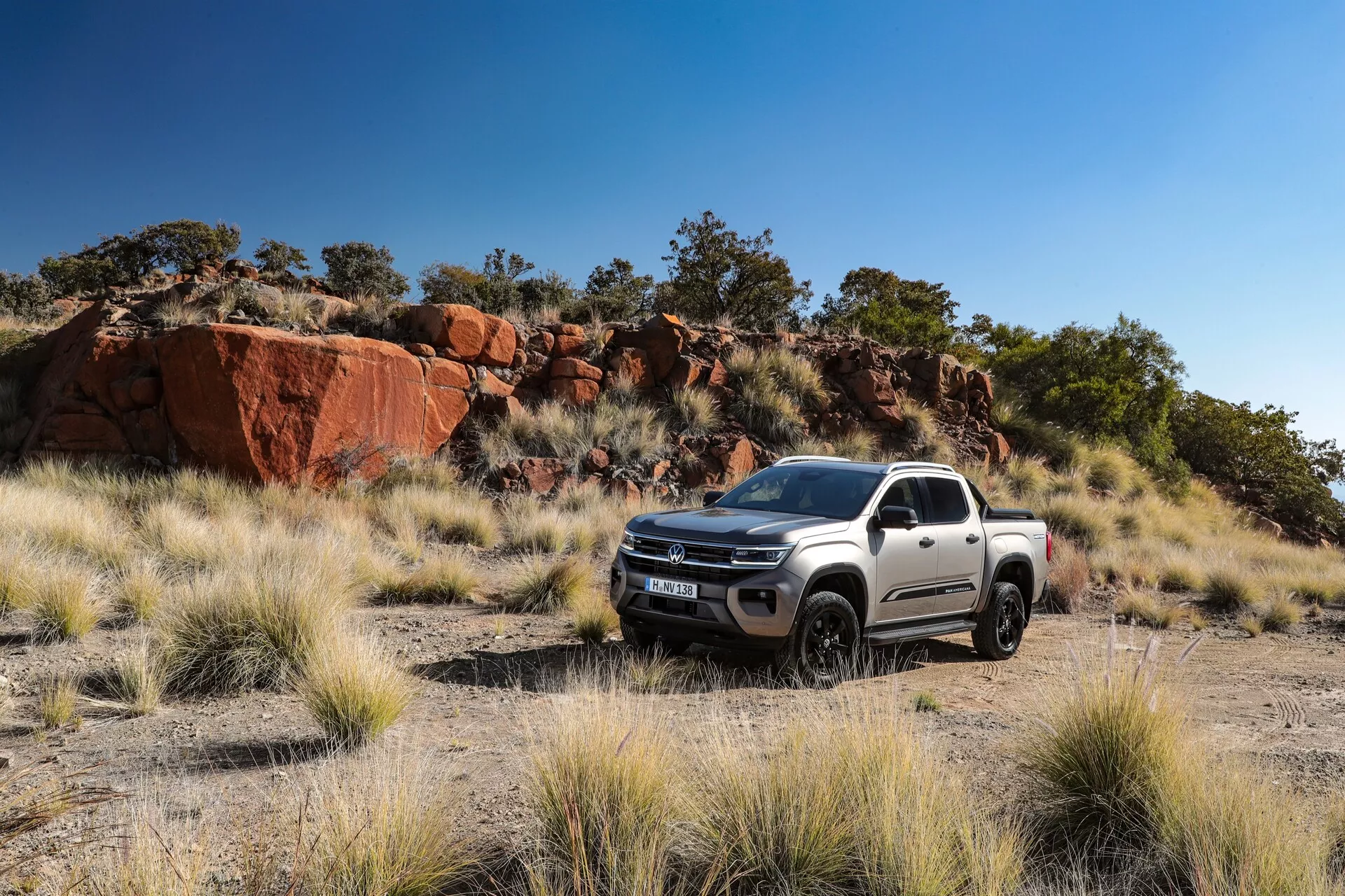 Image d'un pick-up blanc VW-Nutzfahrzeuge Amarok 2023 de deuxième génération dans un cadre extérieur naturel avec une zone herbeuse et un terrain rocheux, soulignant ses capacités tout-terrain. Capturée sous un angle de trois quarts avant, elle montre la grille, les phares, le pare-chocs avant, le capot et les roues du côté conducteur. Le design moderne et les lignes de contour robustes soulignent sa durabilité, avec des jantes en alliage, des composants de suspension visibles et une garde au sol importante. L'éclairage naturel de l'image met en valeur la forme et les caractéristiques du design du véhicule.