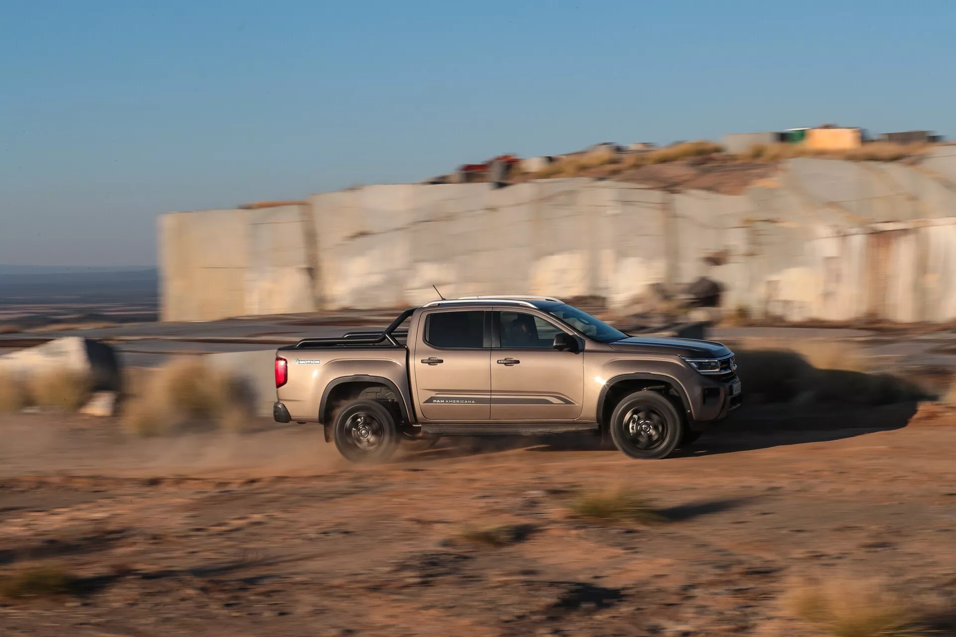 Vue de profil latéral dynamique d'un pickup VW-Nutzfahrzeuge Amarok 2023 (2e génération) en mouvement. Le camion est capturé avec une technique de photographie panoramique, nettement focalisé contre un arrière-plan flou, mettant en avant le mouvement et la vitesse. Il comprend une cabine à double rangée avec quatre portes et des sièges arrière complets, montrant des lignes agressives de la carrosserie, un design moderne des roues, des rétroviseurs latéraux, des poignées de porte et la benne du camion sous une lumière naturelle basse. Cela suggère une utilisation en terrain accidenté extérieur et des technologies automobiles modernes potentielles.