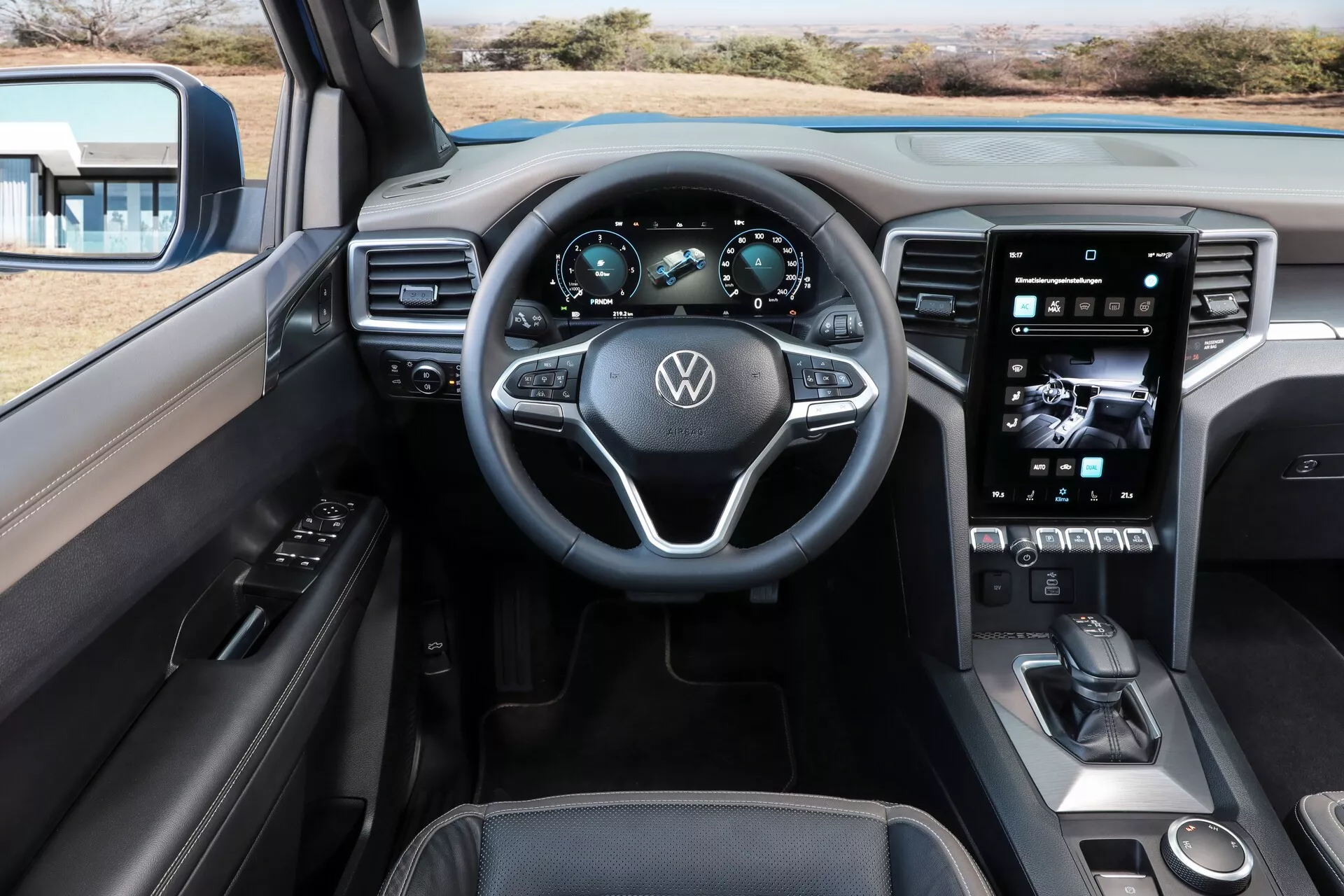 Vue intérieure de la cabine du Volkswagen Amarok 2023 deuxième génération depuis le siège arrière, montrant le volant avec le logo Volkswagen, le tableau de bord numérique, le volant à trois branches avec commandes intégrées, le grand écran tactile vertical du système d'infodivertissement, des commandes physiques minimales suggérant un design axé sur l'écran tactile, un sélecteur de vitesses électronique élégant, un sélecteur de mode de conduite, une possible zone de recharge sans fil pour les téléphones, des matériaux haut de gamme incluant cuir et surfaces souples au toucher avec des coutures détaillées et un éclairage d'ambiance personnalisable.