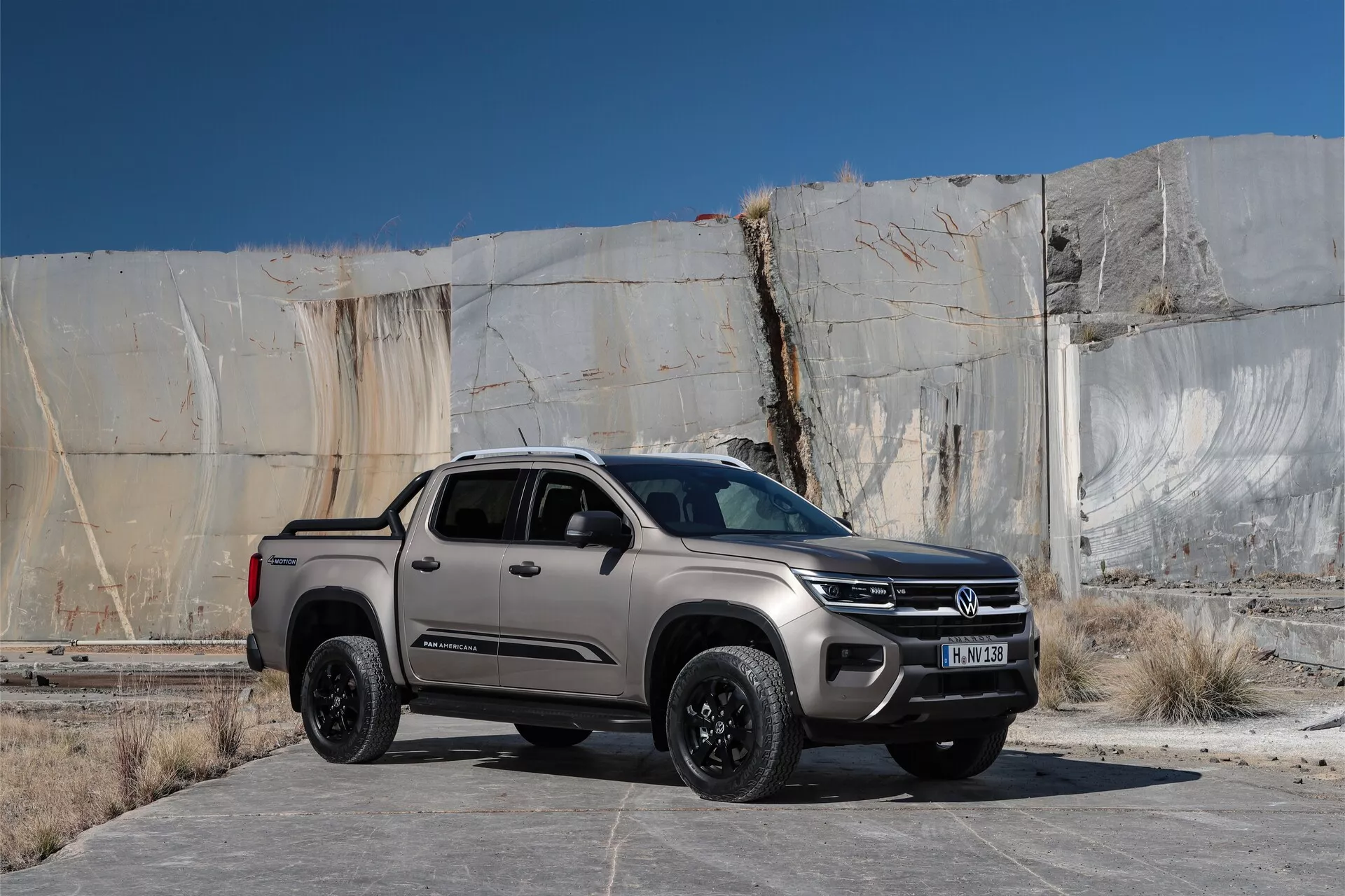 VW Nutzfahrzeuge Amarok 2023 deuxième génération camionnette dans un cadre extérieur, positionnée légèrement à droite du cadre avec un arrière-plan de bâtiment industriel ou abandonné comportant de grandes dalles de béton fissurées. Capturée d'une vue avant trois-quarts à hauteur des yeux, l'image met en évidence la calandre avant, les phares avec des lumières probablement à LED, le capot et le profil latéral, y compris la roue avant côté conducteur et la porte. Le véhicule a une calandre avant proéminente avec un grand badge circulaire, des roues en alliage résistantes noires, une peinture métallique, une garde au sol plus élevée suggérant des capacités 4x4 et des caractéristiques telles que des capteurs de stationnement, des rétroviseurs latéraux avec des clignotants intégrés et une antenne sur le toit. La scène semble être capturée à la lumière du matin ou du pré-soir, mettant en valeur les lignes de design et les ombres.