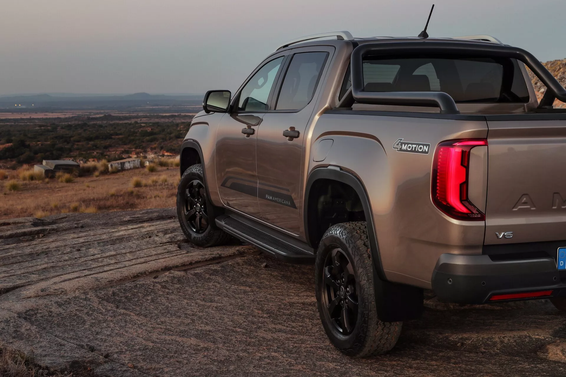 L'image du VW Nutzfahrzeuge Amarok 2023 (2e génération) présente un pick-up sous un angle de trois quarts arrière, mettant en évidence le côté droit et l'arrière. La prise de vue, réalisée d'un angle légèrement en contre-plongée, met en avant la benne du camion, les feux arrière allumés, le pare-chocs arrière et les badges proéminents indiquant la marque, le modèle et peut-être la finition ou les spécifications du moteur. Placé dans un environnement extérieur accidenté avec un terrain visible, le véhicule arbore une finition métallique, une barre de sport dans la benne et des roues noires, suggérant une finition améliorée ou plus sportive. Le badge '4MOTION' indique des capacités de traction intégrale ou à quatre roues, tandis que le logo du hayon et la marche ou poignée intégrée offrent une technologie d'accès facile à la benne.