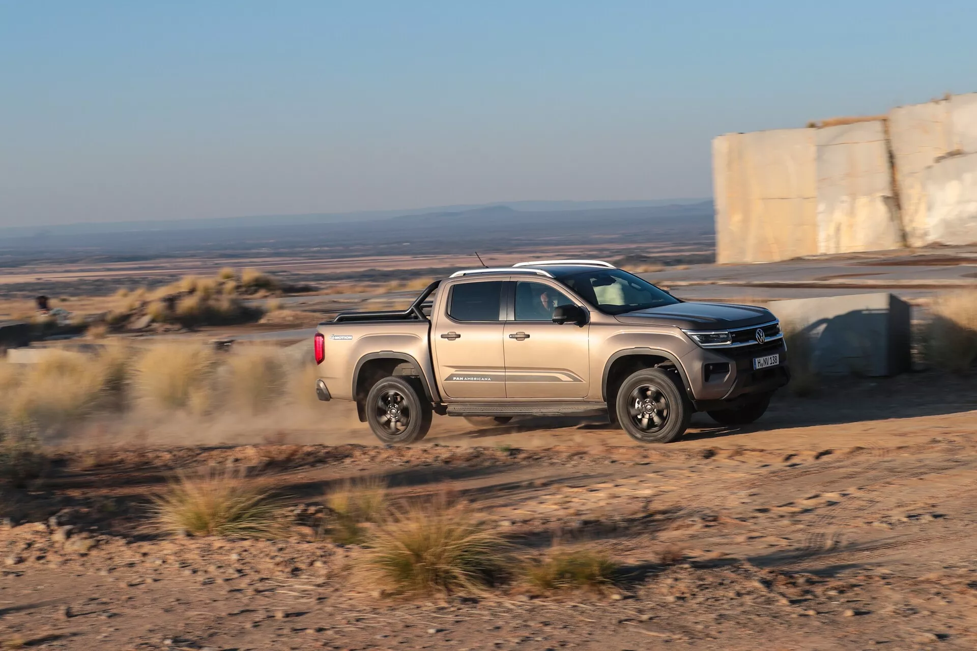 Le pick-up Volkswagen Nutzfahrzeuge Amarok 2023 (2ème génération) capturé en mouvement sur un sentier non pavé, montrant ses capacités tout-terrain avec une traînée de poussière derrière le véhicule. Pris sous un angle oblique, soulignant le côté passager, l'avant et une partie de la benne contre un paysage accidenté en fin d'après-midi.