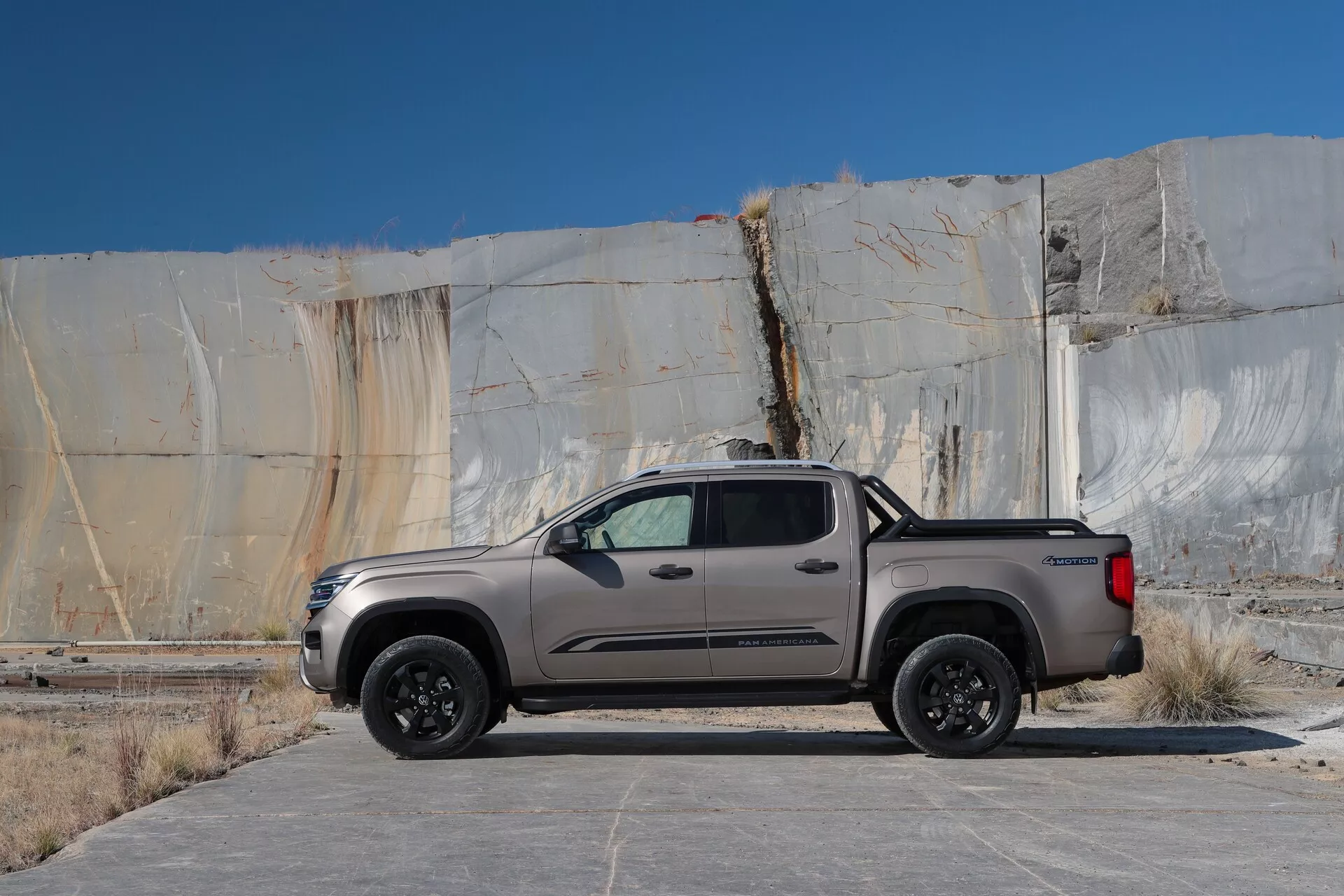 L'image montre un camionnette Volkswagen Amarok 2023 (2ème génération) garée à l'extérieur, positionnée parallèlement à la perspective de la caméra. Le camionnette est une cabine double moderne à quatre portes avec un court compartiment de charge, vue de côté pour révéler toute sa longueur et sa hauteur. Le véhicule est de couleur métallique, soit gris soit brun clair, avec des lignes aérodynamiques et une carrosserie élégante. Il présente des courbes douces qui se rétrécissent vers l'arrière, une ligne de ceinture haute et des vitres teintées à l'arrière. Le camionnette est monté sur des roues noires à multiples rayons, ce qui indique une version améliorée ou un pack sport. Elle est équipée de marchepieds pour faciliter l'accès et de rétroviseurs latéraux proéminents qui peuvent intégrer des indicateurs de signal ou une surveillance des angles morts. Les poignées de porte de la couleur de la carrosserie ajoutent à l'esthétique cohésive. Le compartiment a une couche de protection solide pour prévenir les dommages causés par le chargement, et le hayon arrière présente probablement des technologies comme une caméra de recul. Le camionnette est garé sur une surface en béton ou en asphalte avec quelques taches légères devant une structure avec de grandes dalles en marbre, suggérant un fond industriel ou de carrière. L'éclairage en plein jour avec des ombres minimales indique un ciel nuageux ou un environnement lumineux contrôlé.