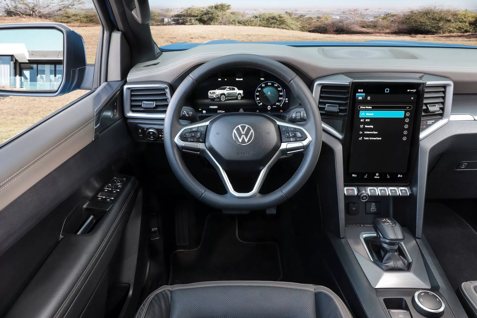 Vue intérieure du Volkswagen Nutzfahrzeuge Amarok 2023 depuis la position centrale des sièges arrière. Les caractéristiques visibles incluent un volant multifonction à trois branches avec logo VW et revêtement en cuir, un tableau de bord numérique, un grand système d'info-divertissement à écran tactile vertical, des commandes physiques minimales, une interface de contrôle climatique élégante et un levier de vitesse moderne avec frein de stationnement électronique. Le panneau de porte du conducteur est partiellement visible, avec des commandes de fenêtres et de rétroviseurs latéraux. L'intérieur présente une combinaison de matériaux premium, comprenant du cuir, des plastiques souples et des accents métalliques dans une gamme de couleurs sombre à deux tons. Les sièges avant sont visibles avec des appuie-tête intégrés et des surpiqûres contrastées, indicatives d'une finition luxueuse.