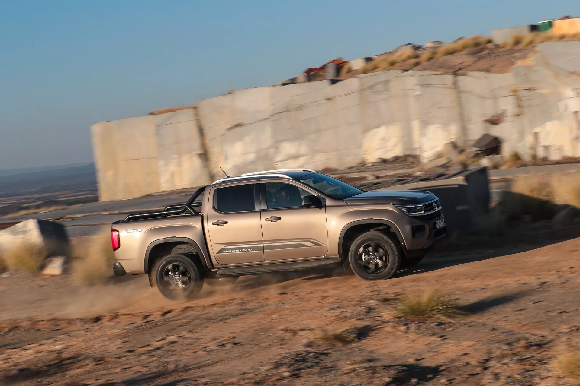 Une photo de profil du pickup VW Nutzfahrzeuge Amarok 2023 de deuxième génération en mouvement. Le camion se déplace de gauche à droite à travers le cadre dans un environnement tout-terrain, soulevant de la poussière derrière les roues arrière. La lumière chaude et basse du soleil souligne le profil du véhicule, y compris ses roues robustes, ses phares LED modernes et sa grille avant distinctive.