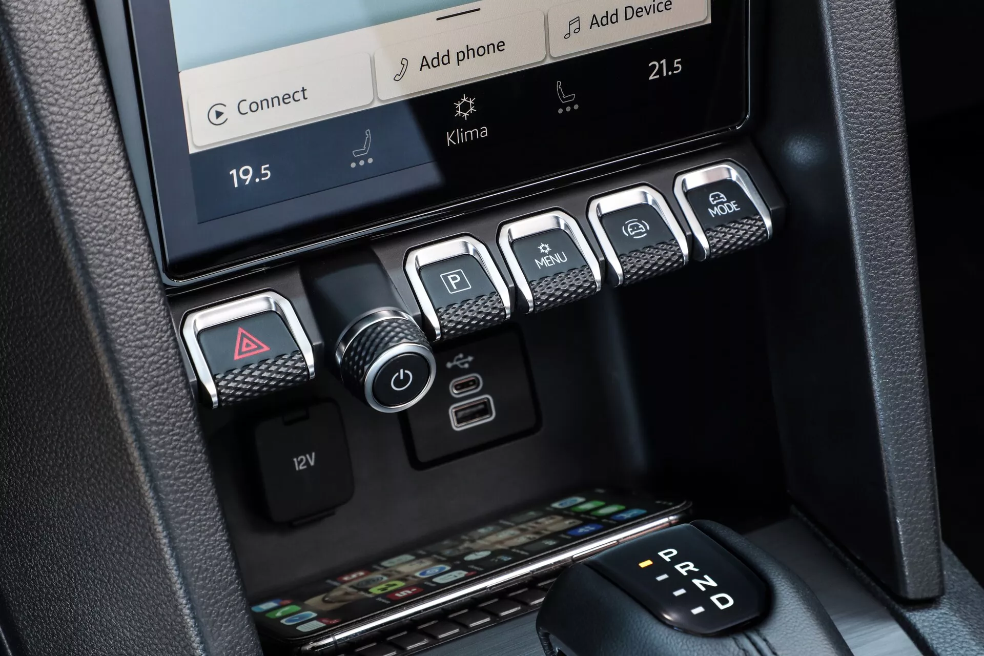 Gros plan de la console centrale du VW Nutzfahrzeuge Amarok 2023 de 2e génération, montrant le contrôle du climat bi-zone avec écran tactile, affichant des températures de 19,5°C et 21,5°C. Le bord du système d'infodivertissement est visible au-dessus, proposant des options de connectivité telles que 'Connect' et 'Ajouter Téléphone.' En dessous, cinq boutons métalliques pour les feux de détresse, les capteurs de stationnement, le démarrage/arrêt du moteur, les modes de conduite ou contrôle de traction, et le contrôle de descente, adjacents à une prise de courant 12V. En bas, un compartiment de rangement pour clés et portefeuilles. Le côté droit du cadre révèle le sélecteur de transmission automatique affichant 'P' pour le stationnement. Les matériaux et finitions dégagent un design premium et moderne.