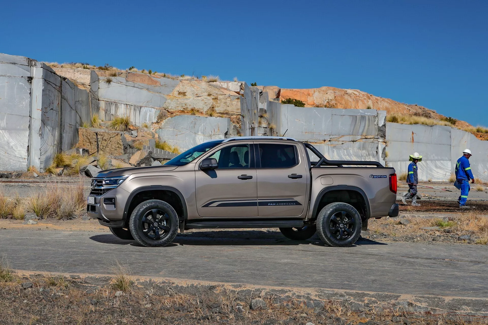 L'image montre une VW Nutzfahrzeuge Amarok 2023 de deuxième génération, un pickup à quatre portes positionné en extérieur avec son profil latéral face à la caméra. Le véhicule est photographié depuis un angle légèrement bas, mettant en valeur sa garde au sol et sa stature robuste, idéales pour des terrains variés. Le camion est stationné parallèlement à la perspective de la caméra, offrant une vue claire de son côté, qui comprend les portes avant et arrière passagers, les rétroviseurs extérieurs, les poignées, les lignes de design, les passages de roues et les roues. Le véhicule est situé sur une surface plane non pavée qui semble être un chantier de construction, avec un terrain poussiéreux et des travailleurs en arrière-plan, soutenant son adéquation pour une utilisation industrielle ou commerciale. Les points focaux de l'image incluent les capacités tout-terrain, le design de la suspension, le forfait roues et pneus, ainsi que la finition de la peinture et le niveau de finition du véhicule.