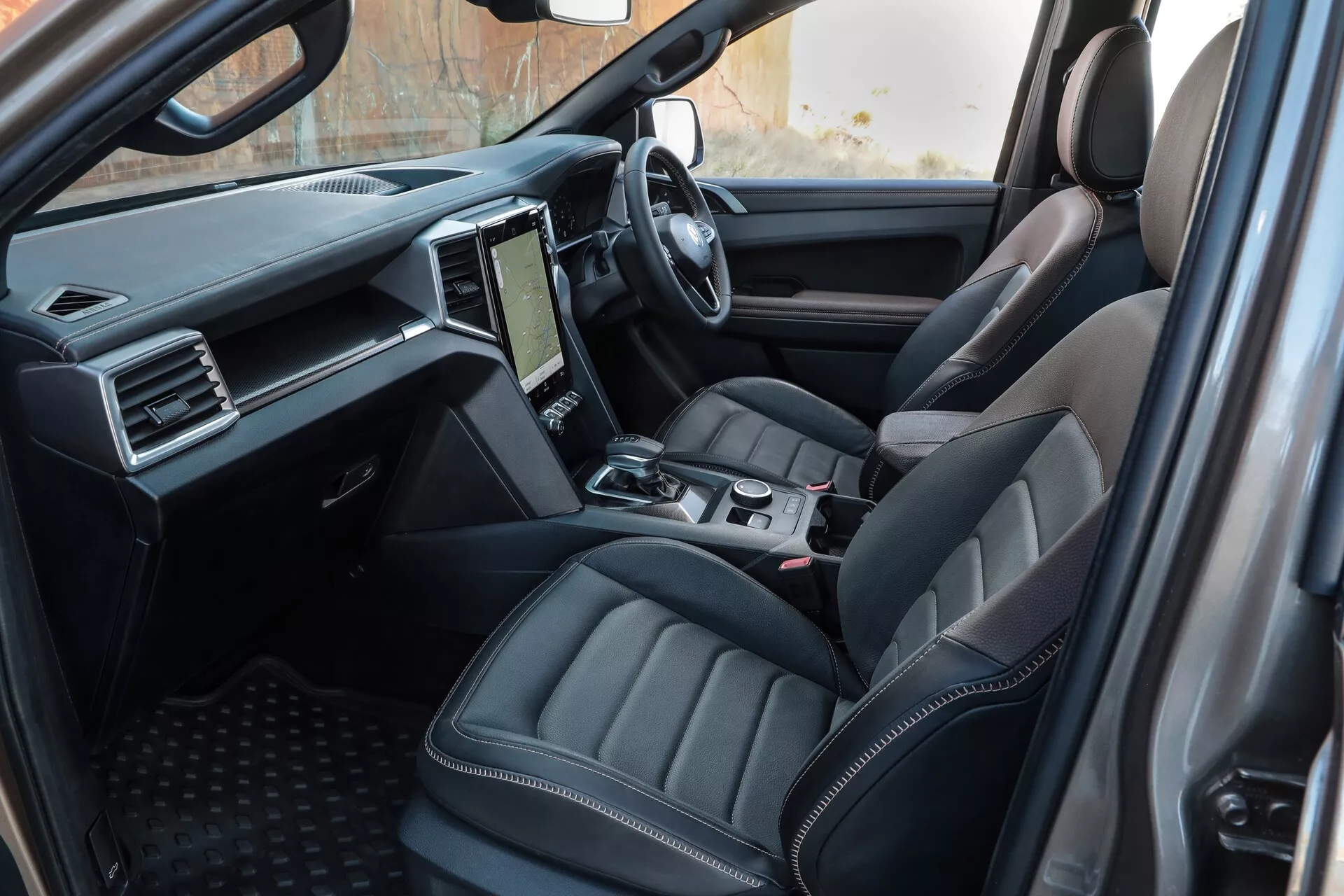 Vue intérieure du VW Nutzfahrzeuge Amarok 2023 (2e génération), capturée de la perspective d’un passager arrière, mettant en évidence le siège du conducteur en cuir avec des contours, la console centrale avec commandes et le tableau de bord moderne avec système d'infodivertissement à écran tactile.