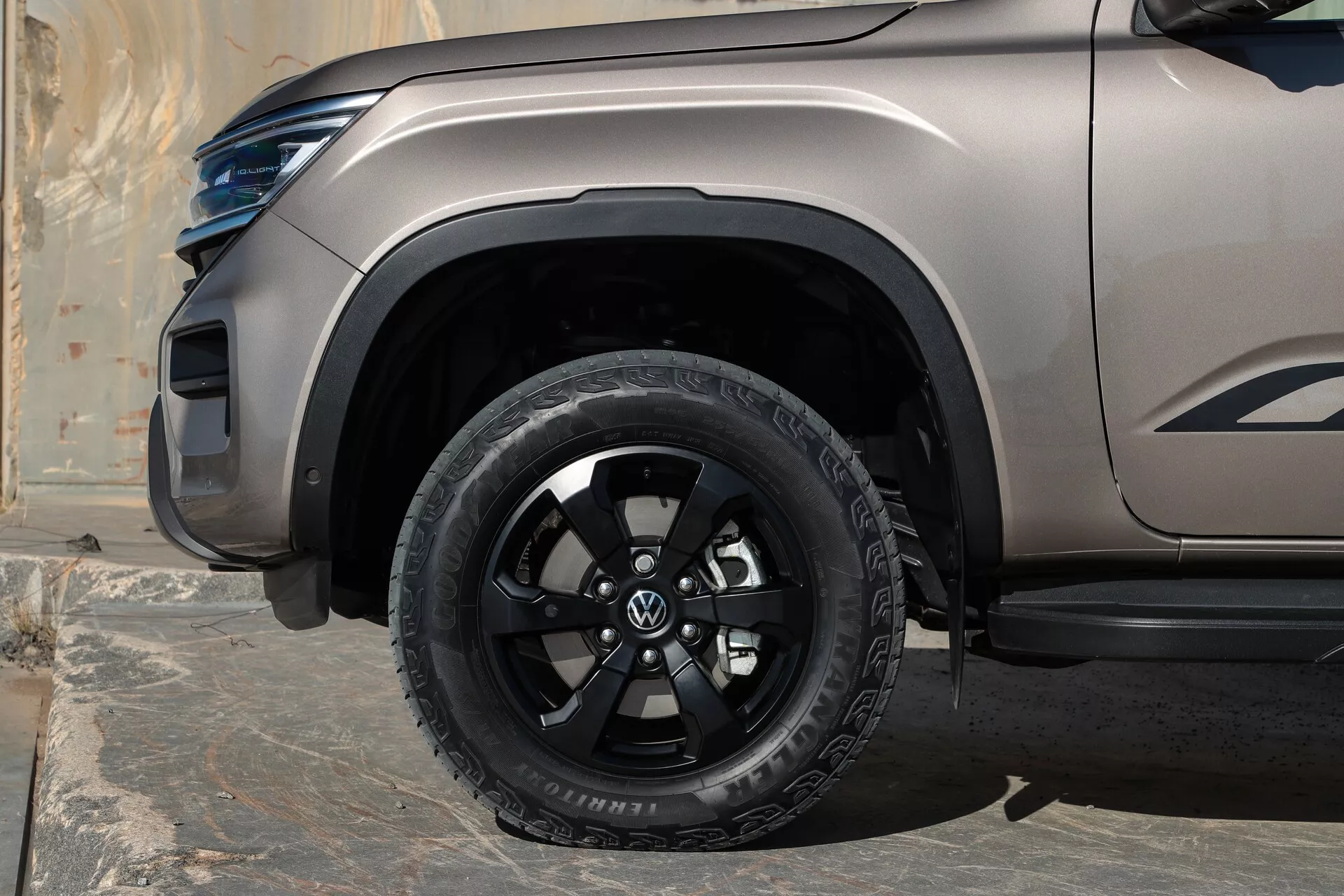 Vue rapprochée de la roue avant du Volkswagen Amarok 2023 (2ème génération), avec une roue à multiples rayons noire brillante et un pneu tout-terrain, une finition de peinture mate/satinée, un élargisseur d'aile en plastique, une marchepied sous la porte passager, et un phare LED et une ligne de carrosserie sculptée partiellement visibles.