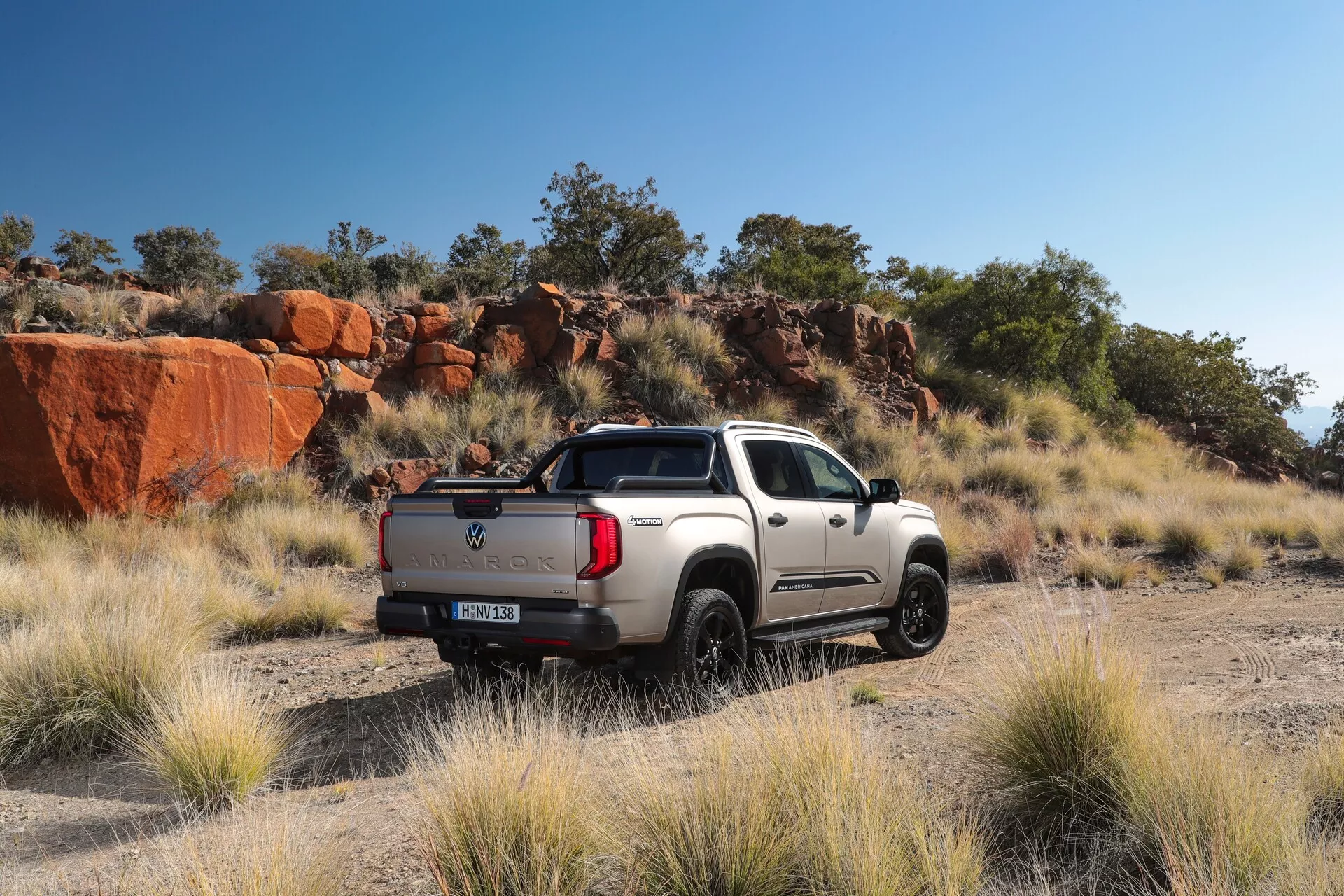 Image d'un pick-up Volkswagen Nutzfahrzeuge Amarok 2023 garé en plein air sur un chemin de terre avec un paysage rocheux naturel en arrière-plan, capturé d'une perspective arrière trois quarts. Le camion élégant avec des accents chromés est illuminé par la lumière naturelle, mettant en valeur son design sportif mais robuste avec une configuration double cabine, un lit court, des ailes proéminentes et des pneus à bande de roulement épaisse adaptés aux aventures tout-terrain.