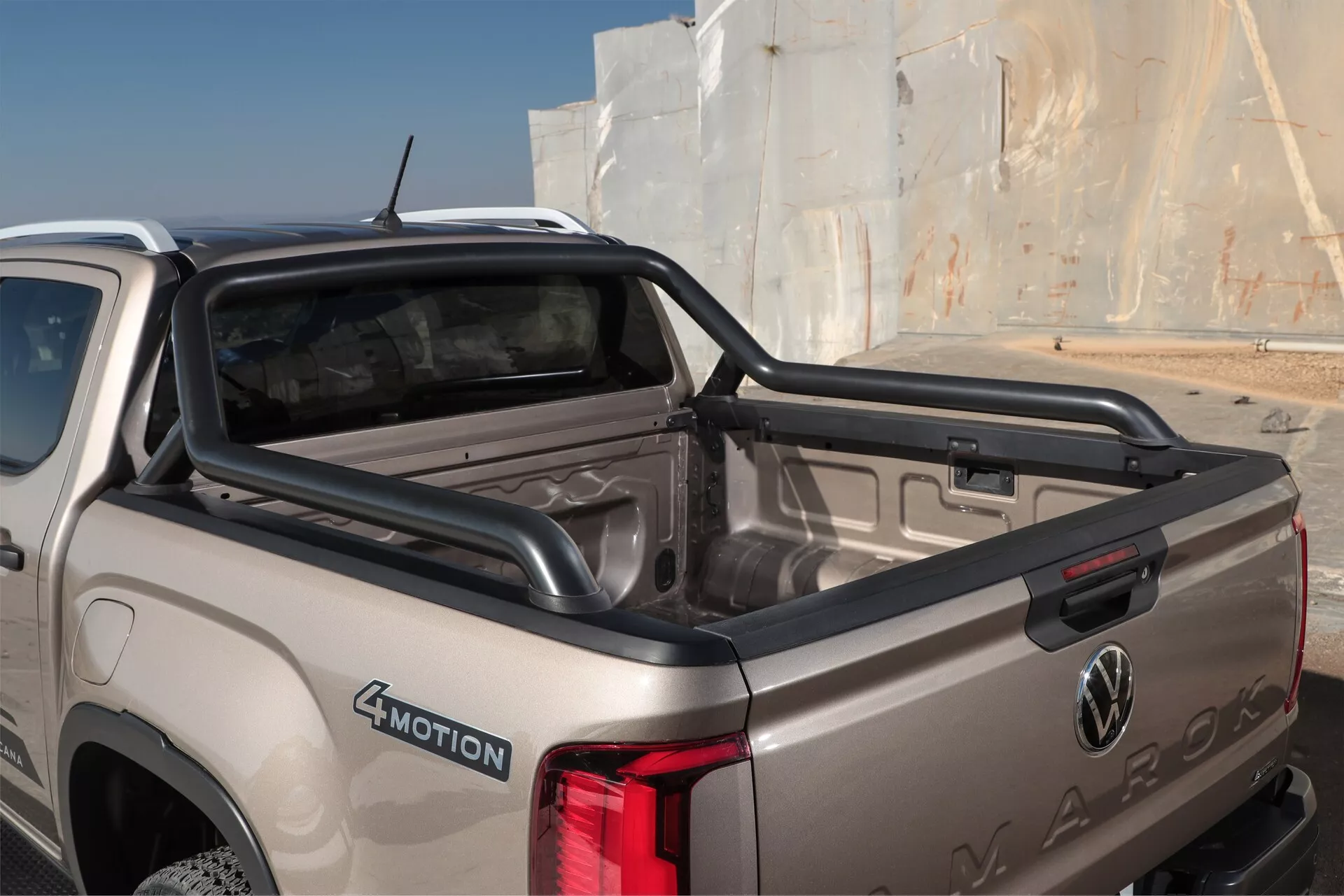 Vue rapprochée et en angle de la section arrière d'un pick-up Volkswagen Amarok 2023 (2e génération) beige ou or clair. Le cliché se concentre sur la zone du lit et du hayon, mettant en avant le logo Volkswagen et le nom Amarok estampillé dans le métal. Les caractéristiques visibles incluent un lit court avec doublure en plastique, l'ensemble de feux arrière côté droit, l'insigne 4MOTION de la transmission intégrale et une marche de pare-chocs pour un accès plus facile. La fenêtre arrière, potentiellement dotée d'un dégivreur, offre une vue sur l'habitacle propre et bien entretenu. L'image capture les lignes sculptées de la carrosserie, indiquant un design automobile sophistiqué en lumière naturelle diurne.