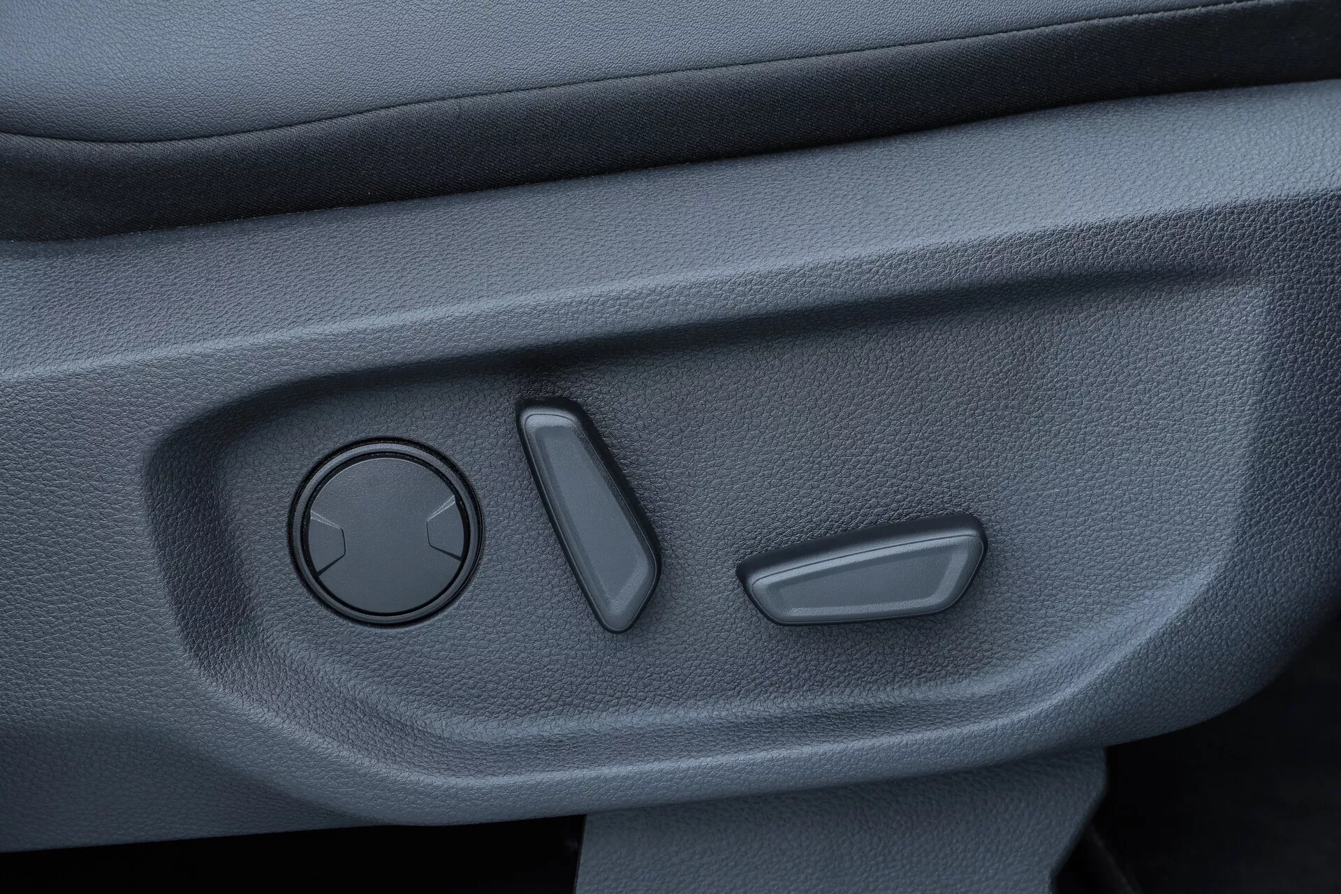 Gros plan du panneau de porte intérieur du VW Nutzfahrzeuge Amarok 2023, présentant un bouton rond pour le réglage des rétroviseurs, deux interrupteurs alignés verticalement probablement pour les vitres électriques et le contrôle de verrouillage des portes, et une surface texturée de couleur sombre indiquant un matériau durable et antidérapant. La photographie est prise sous un léger angle vers le bas pour afficher clairement les commandes.