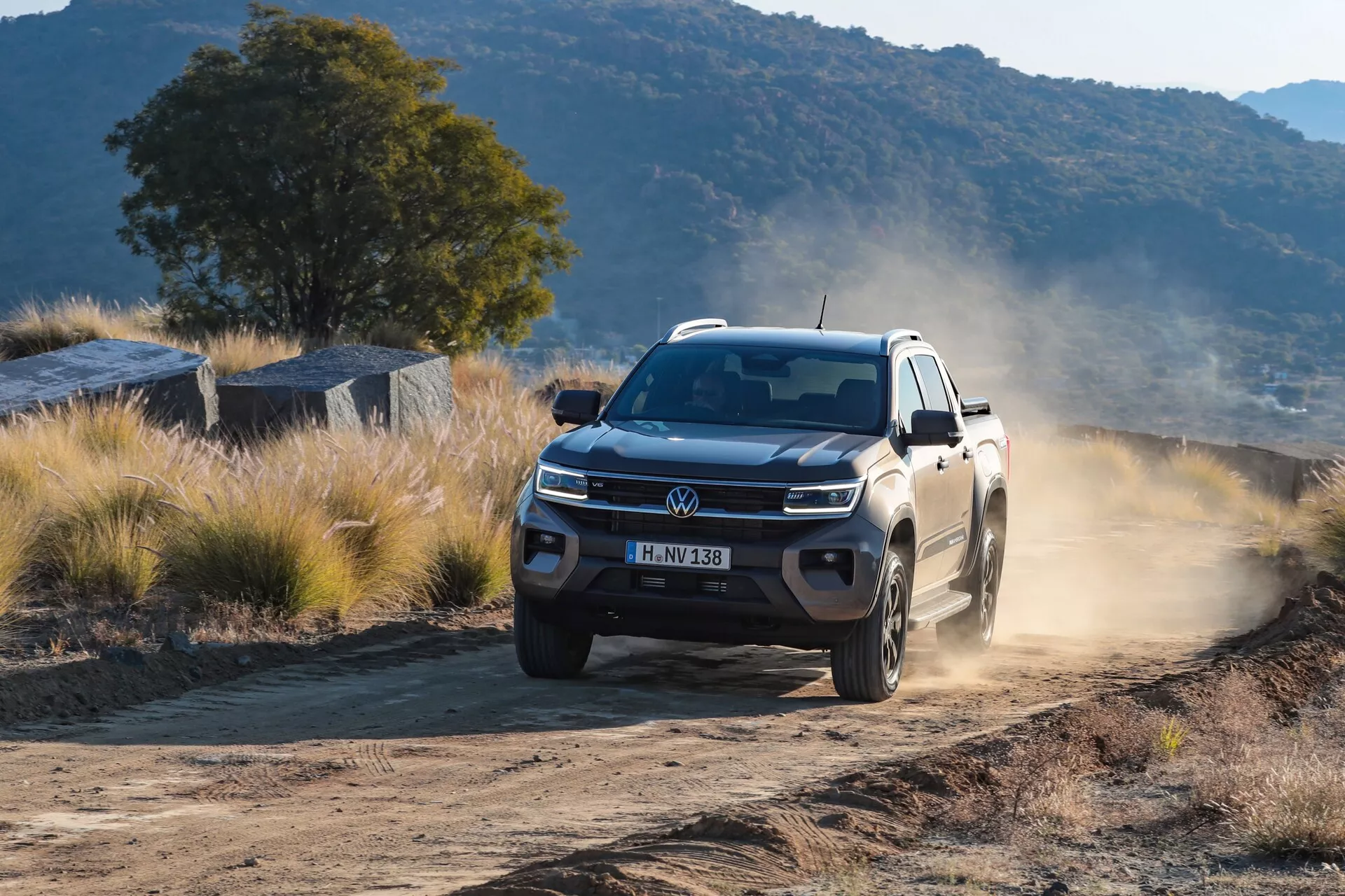 Photographie professionnelle d'un pick-up Volkswagen Nutzfahrzeuge Amarok 2023 de deuxième génération, capturée sous un angle de trois quarts avant. L'image met en avant la face avant moderne et robuste du véhicule, la grille proéminente avec le logo Volkswagen et les feux de jour à LED. L'Amarok est en mouvement sur une route poussiéreuse ou de gravier dans un cadre naturel, soulevant de la poussière des roues arrière, signifiant ses capacités tout-terrain et son utilité en dehors des routes. Les roues en alliage, la garde au sol élevée et la suspension contribuent aux performances et à la durabilité du camion, pertinent pour la catégorie SUV/tout-terrain. L'angle bas de la prise de vue souligne la présence dominante et puissante du véhicule.