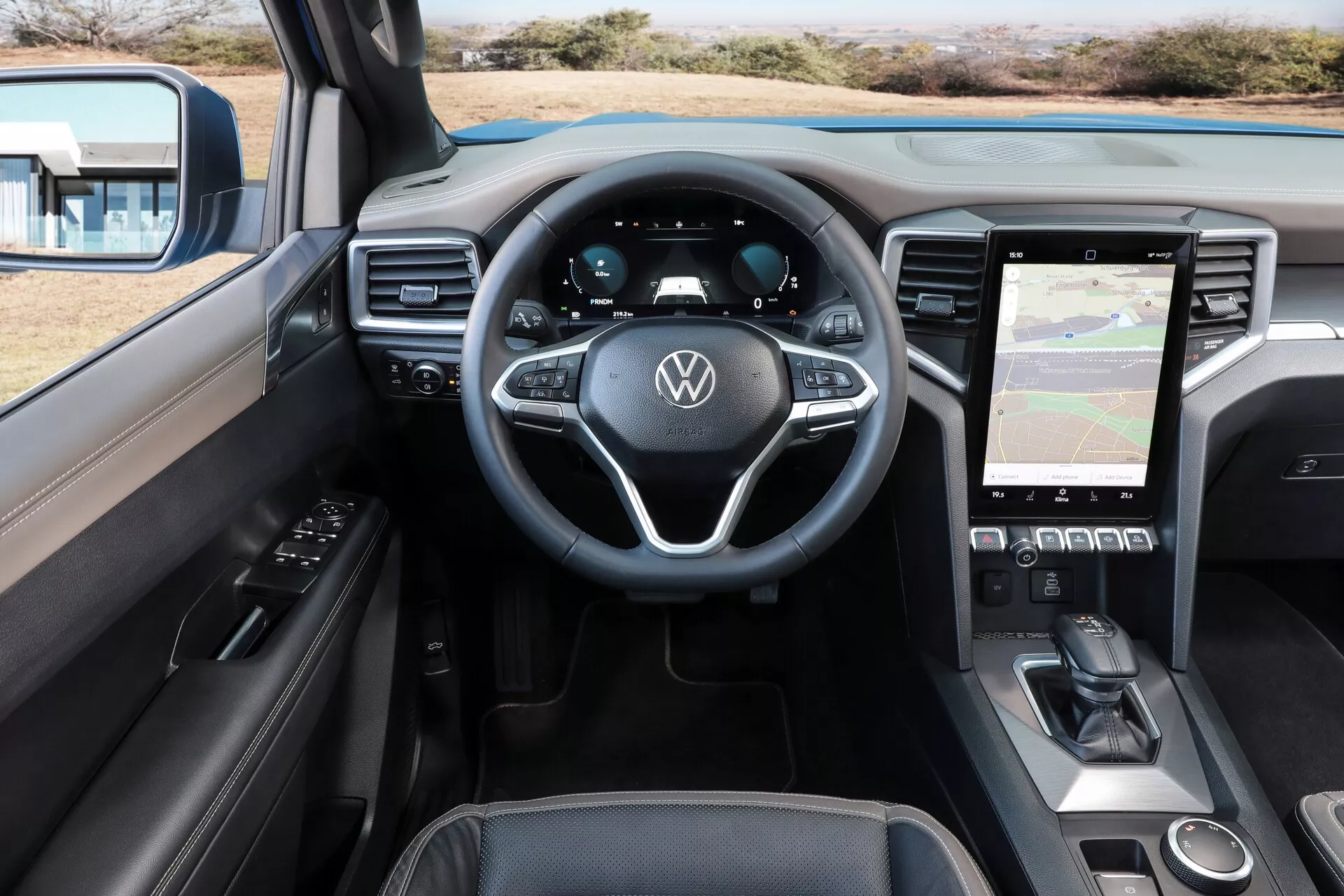 Vue de l'intérieur de la cabine du VW Nutzfahrzeuge Amarok 2023 (2e génération) depuis la perspective du conducteur. L'image est prise sous un angle élevé, montrant l'avant de la cabine. Dans le cadre, on peut voir le volant avec le logo Volkswagen et des commandes multifonctions, un tableau de bord entièrement numérique et un système d'infodivertissement à écran tactile vertical affichant une carte. La console centrale comprend des commandes de climatisation, une zone potentielle de recharge sans fil et un levier de vitesse automatique. Le design élégant et contemporain présente des matériaux en cuir et doux au toucher sur le tableau de bord et les panneaux de porte, avec des boutons de commande des fenêtres et des ajustements des miroirs sur la porte du conducteur.