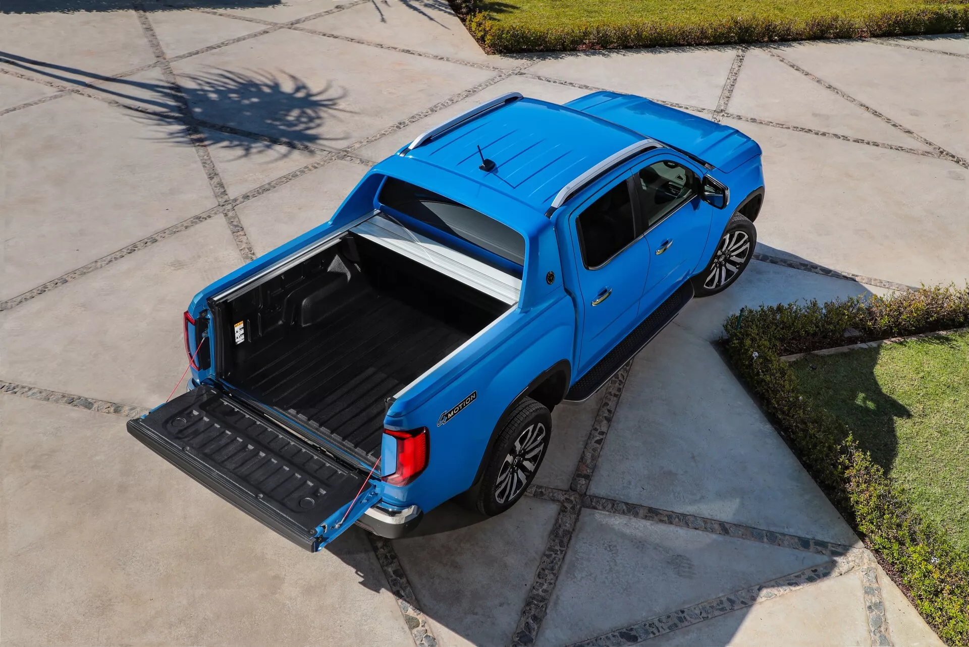 Vue aérienne d'un pick-up bleu VW Nutzfahrzeuge Amarok seconde génération de 2023. L'image montre le véhicule avec une cabine double, des sièges arrière de taille normale et quatre portes. La benne est ouverte et vide, avec le hayon abaissé, soulignant l'utilité du véhicule. Les lignes élégantes de la carrosserie, les jantes en alliage dans les arches de roues visibles et les poignées de portes de couleur carrosserie sont visibles. L'environnement semble résidentiel avec des trottoirs bien entretenus, sous un ciel dégagé avec le soleil haut.