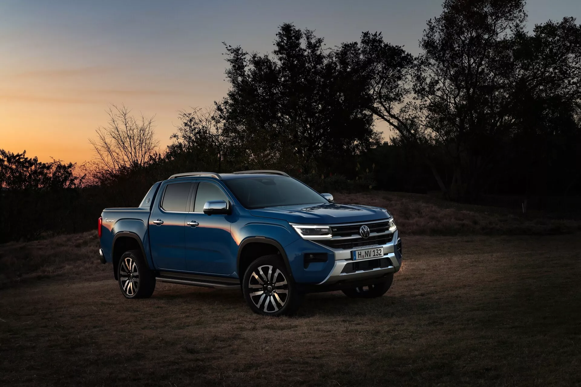 Profil latéral d'un pickup bleu VW Nutzfahrzeuge Amarok 2023 (2e génération) au crépuscule ou à l'aube, montrant le côté du véhicule, la calandre avant et les phares avec un horizon teinté d'orange et une ambiance de faible luminosité. Les caractéristiques de conception du véhicule, y compris les phares LED modernes, la calandre avant chromée, les clignotants intégrés dans les rétroviseurs latéraux, le design des roues en alliage et la finition réfléchissante de la peinture, sont mises en valeur par un éclairage artificiel focalisé. La scène équilibre le caractère aventurier et robuste du pickup avec son environnement naturel en extérieur, soulignant sa polyvalence et ses capacités tout-terrain.
