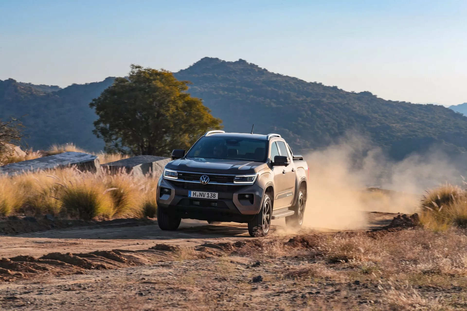 L'image montre un pick-up VW Nutzfahrzeuge Amarok 2023 de deuxième génération en train de rouler sur une route non pavée, probablement en terre ou en gravier, dans un paysage rural ou montagneux. Le véhicule est photographié sous un angle trois-quarts avant, montrant ainsi sa calandre, son pare-chocs, ses phares, ses feux antibrouillard, son capot, ses ailes avant, ses roues et son profil latéral. L'éclairage doux, probablement en matinée ou fin d'après-midi, met en évidence le nuage de poussière soulevé par le véhicule en mouvement, indiquant sa vitesse et les conditions de sécheresse du terrain. Les éléments de design du véhicule, tels que les roues adaptées aux tout-terrains, le pare-chocs avant robuste et l'éclairage avancé, suggèrent des caractéristiques et des technologies modernes, incluant le contrôle de traction, la stabilité en conditions difficiles et éventuellement des systèmes avancés d'assistance au conducteur.