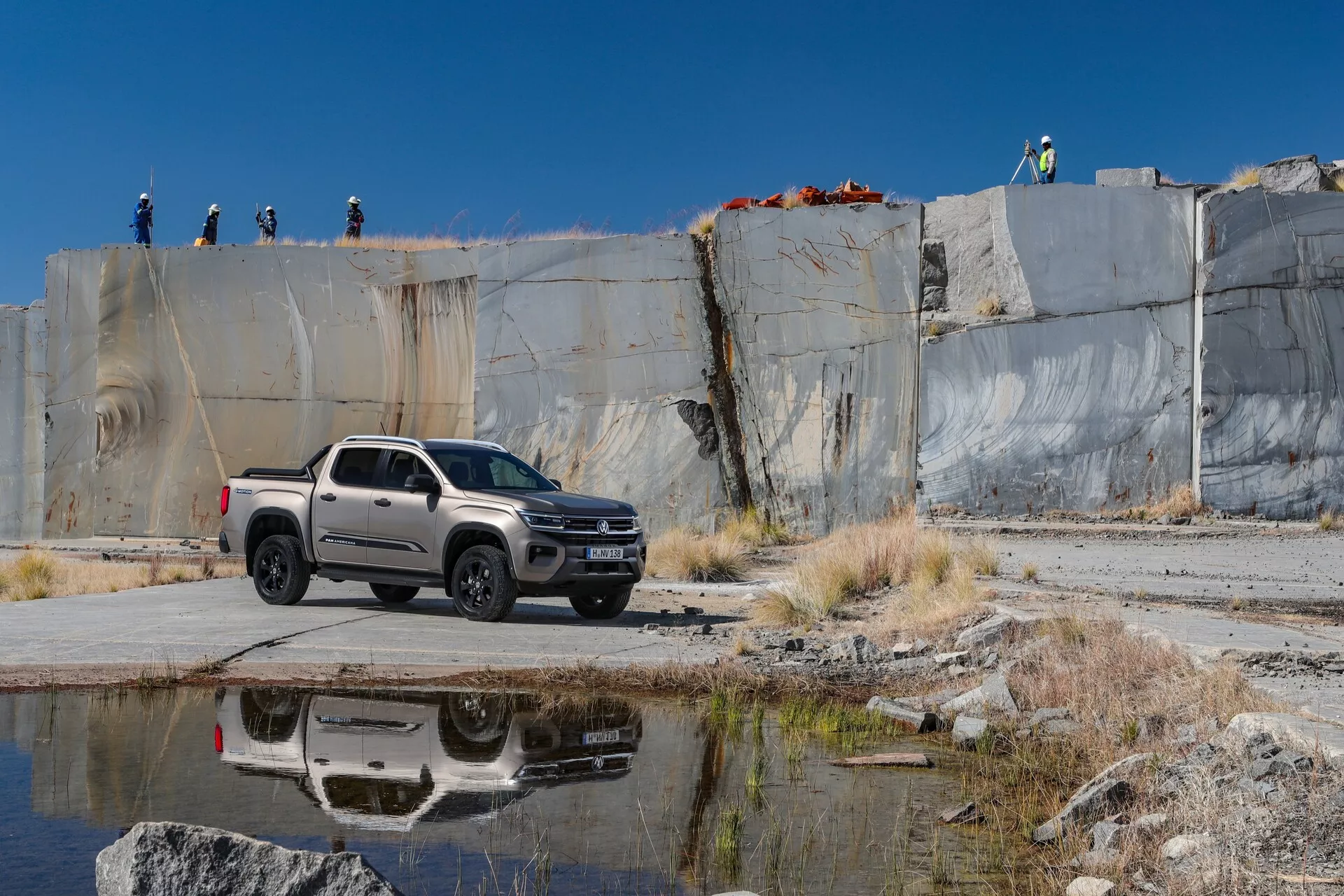 L'image montre un pick-up VW Nutzfahrzeuge Amarok 2023 de deuxième génération garé sur une surface en béton ou pavée dans une carrière ou un chantier de construction, vu sous un angle frontal trois-quarts. La face avant du véhicule, y compris les phares, la calandre et le pare-chocs, ainsi que son profil latéral avec des roues, des portes et des rétroviseurs latéraux, sont clairement visibles. Le camion présente un design moderne avec des lignes de carrosserie robustes, une configuration en cabine double et un plateau court, peint dans une couleur claire, possiblement argentée ou grise. La lumière naturelle met en évidence les caractéristiques de conception et la peinture. En arrière-plan, des personnes portant des casques de sécurité sont visibles sur une corniche surélevée, ce qui suggère un chantier industriel ou de construction actif. Les caractéristiques technologiques visibles incluent des jantes en alliage, des rétroviseurs latéraux avec des clignotants intégrés et des phares à LED. Le reflet du camion sur un plan d'eau voisin ajoute une dimension esthétique et suggère ses capacités tout-terrain.
