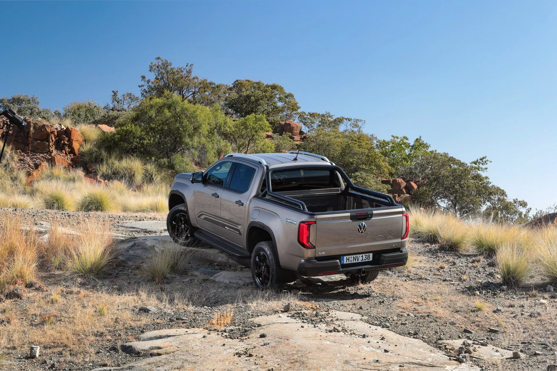 Pick-up Volkswagen Amarok 2023 de deuxième génération dans un environnement tout-terrain, vue arrière trois-quarts. Le véhicule est garé sur un chemin de terre irrégulier, avec des broussailles naturelles et une colline rocheuse en arrière-plan. Les caractéristiques visibles comprennent le hayon avec l'emblème du constructeur, le pare-chocs arrière robuste, les roues en alliage avec des pneus tout-terrain, le lit de chargement accessible, les feux arrière proéminents, le bouchon de carburant sur le panneau arrière et les arches de garde-boue robustes.