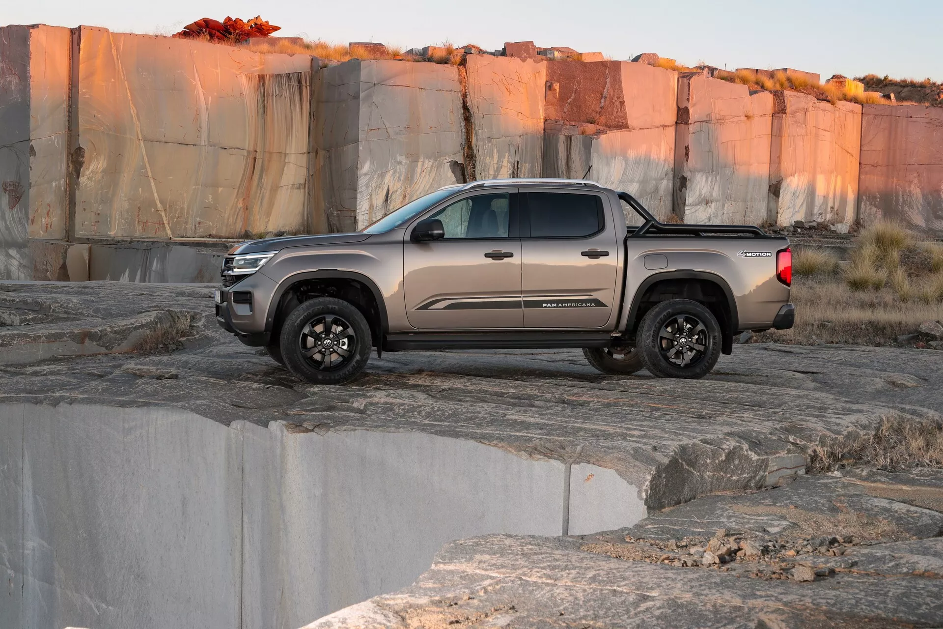 Vue latérale d'un VW Nutzfahrzeuge Amarok 2023, deuxième génération, un pick-up de taille moyenne avec une configuration cabine double et des pneus tout-terrain, positionné sur un terrain rocailleux et accidenté suggérant une carrière ou un environnement naturel au lever ou au coucher du soleil. La prise de vue en contre-plongée met en valeur la garde au sol et la robustesse du camion. Le véhicule est de couleur taupe ou grise atténuée avec des roues à finition foncée, capturant l'essence du design automobile moderne.