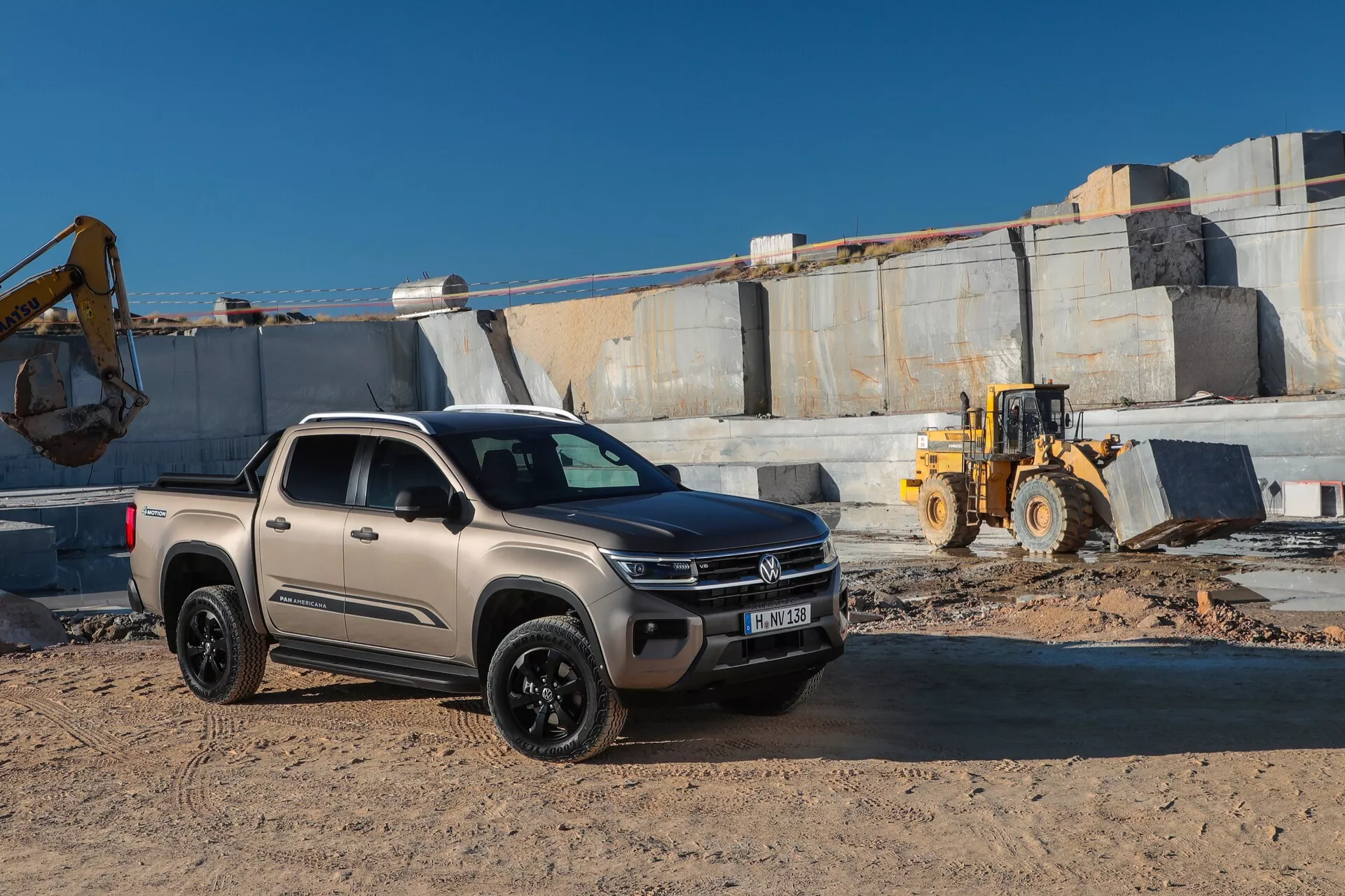 Le pick-up VW Nutzfahrzeuge Amarok 2023 de deuxième génération est garé sur un chantier avec des éléments de carrière en arrière-plan. La photo est prise sous un angle bas, montrant la grille avant, les phares, le capot et le profil latéral côté passager. Il présente une configuration double cabine, un design moderne avec des lignes élégantes, une apparence robuste avec des pneus tout-terrain et une garde au sol accrue. La peinture du camion est mate, probablement beige ou taupe clair, avec des roues noires, des phares LED avec feux de jour. Le cadre comprend des machines lourdes et un sol poussiéreux et inégal.