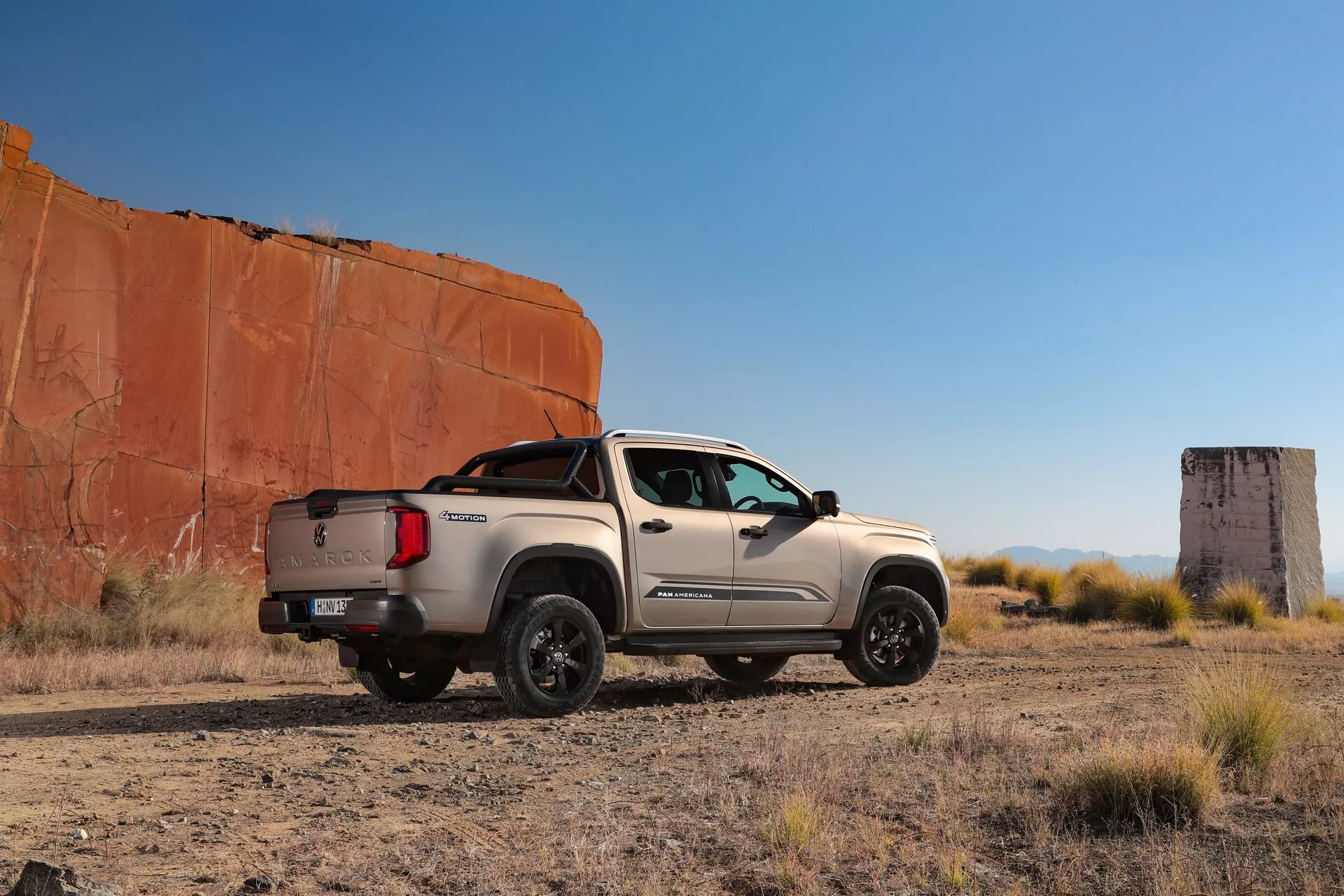Vue latérale d'un pick-up VW Nutzfahrzeuge Amarok 2023 capturé sous un angle arrière légèrement incliné, stationné sur un terrain sec et plat dans un environnement désertique avec une végétation clairsemée et un ciel bleu clair, avec en arrière-plan de grandes formations rocheuses rouges. Le profil latéral du camion, y compris la calandre avant, les phares, la porte côté conducteur, le profil côté passager, la benne et le hayon, sont entièrement visibles. Des caractéristiques distinctives comme les rétroviseurs latéraux, le design des roues et le marchepied contribuent à son aspect robuste. L'angle bas de la prise de vue met en valeur la garde au sol et l'allure puissante, avec des ombres suggérant un éclairage de fin d'après-midi.