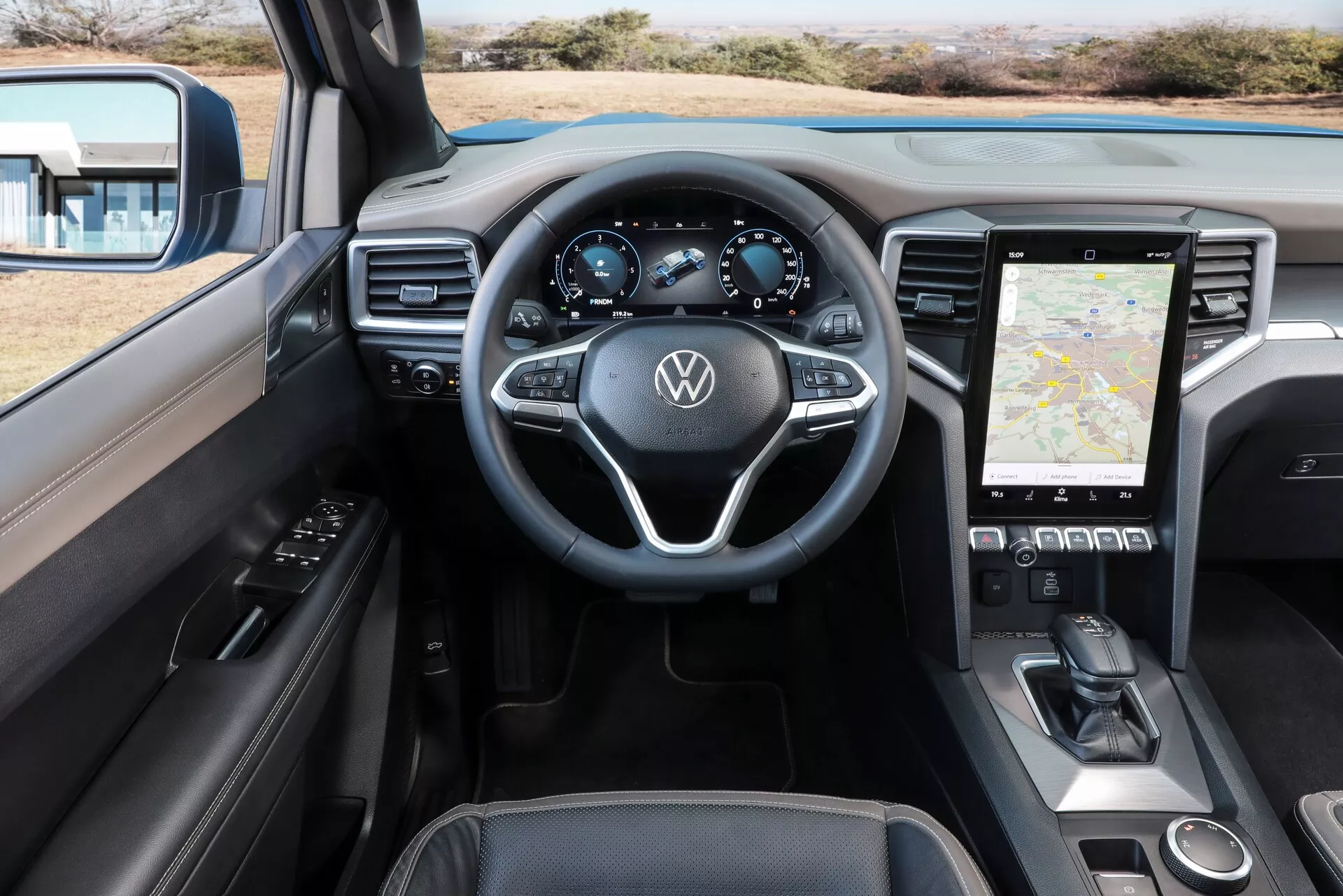 Vue intérieure du Volkswagen Nutzfahrzeuge Amarok 2023, montrant la perspective du conducteur avec vue sur le tableau de bord, le volant et la console centrale. Le volant possède un design à trois branches avec commandes multifonctions et logo Volkswagen. Le tableau de bord numérique du conducteur affiche le compteur de vitesse, le compte-tours, l'indicateur de carburant et possiblement des informations de navigation ou de trajet. La console centrale comprend un grand écran tactile affichant une carte de navigation, avec des commandes de climatisation tactiles en dessous. L'intérieur suggère des technologies embarquées avancées et un design contemporain avec des surfaces douces, des accents métalliques et des garnitures en cuir.