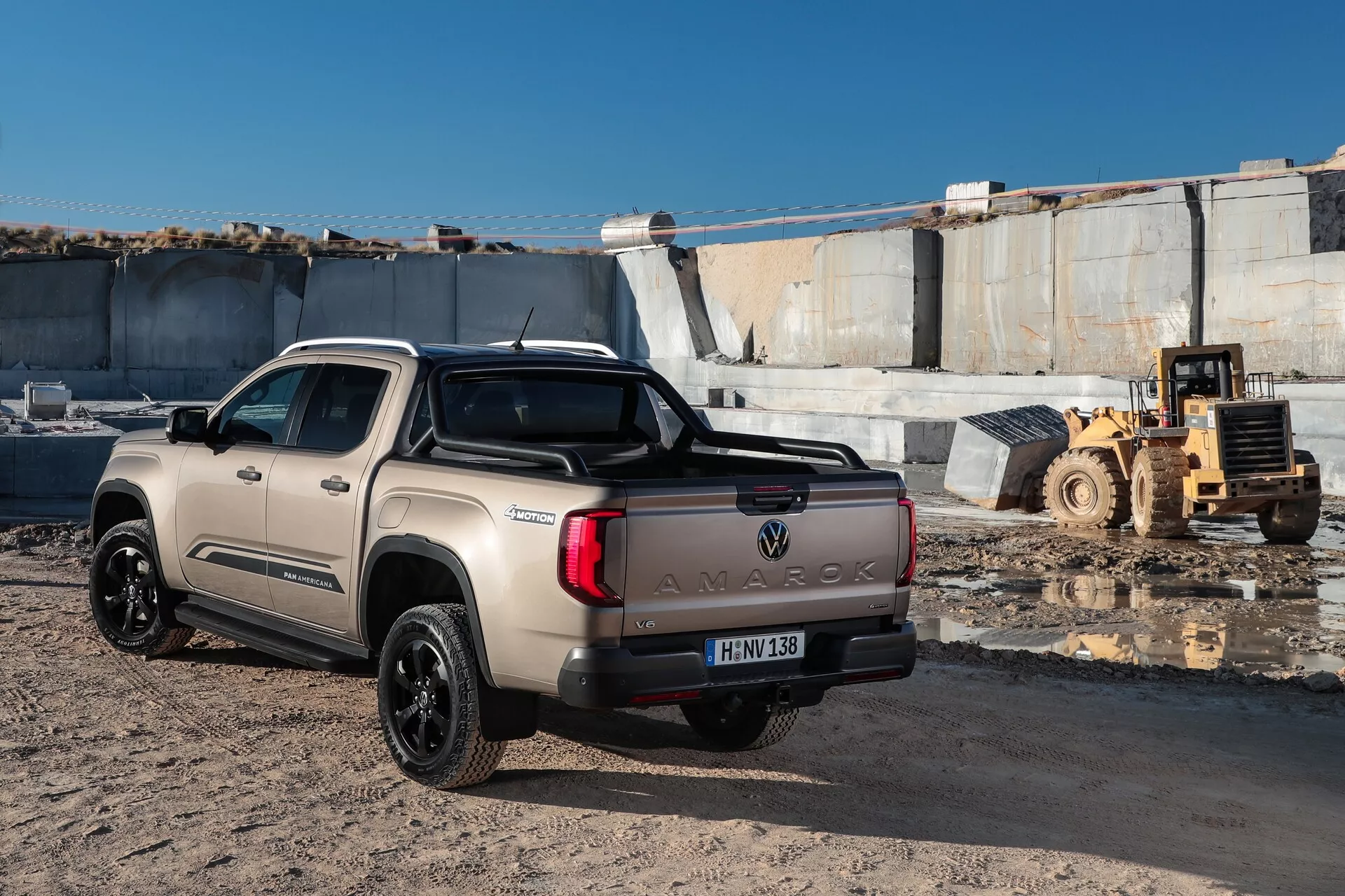 Cette image montre un pick-up Volkswagen Amarok 2023 (deuxième génération) pris sous un angle arrière trois-quarts, mettant en avant le côté droit du véhicule. Le camion est garé sur une surface graveleuse dans un cadre extérieur ensoleillé, probablement dans une zone de construction ou industrielle, comme en témoigne la présence de machines lourdes en arrière-plan. Les caractéristiques principales incluent le lettrage 'AMAROK' en lettres majuscules sur le hayon, une barre de sport sur le lit, des feux arrière horizontaux et des jantes en alliage sombre à plusieurs branches. La peinture beige clair et la configuration de la cabine double avec quatre portes complètes suggèrent une cabine spacieuse. La plaque d'immatriculation dans le coin inférieur gauche est floue pour des raisons de confidentialité.