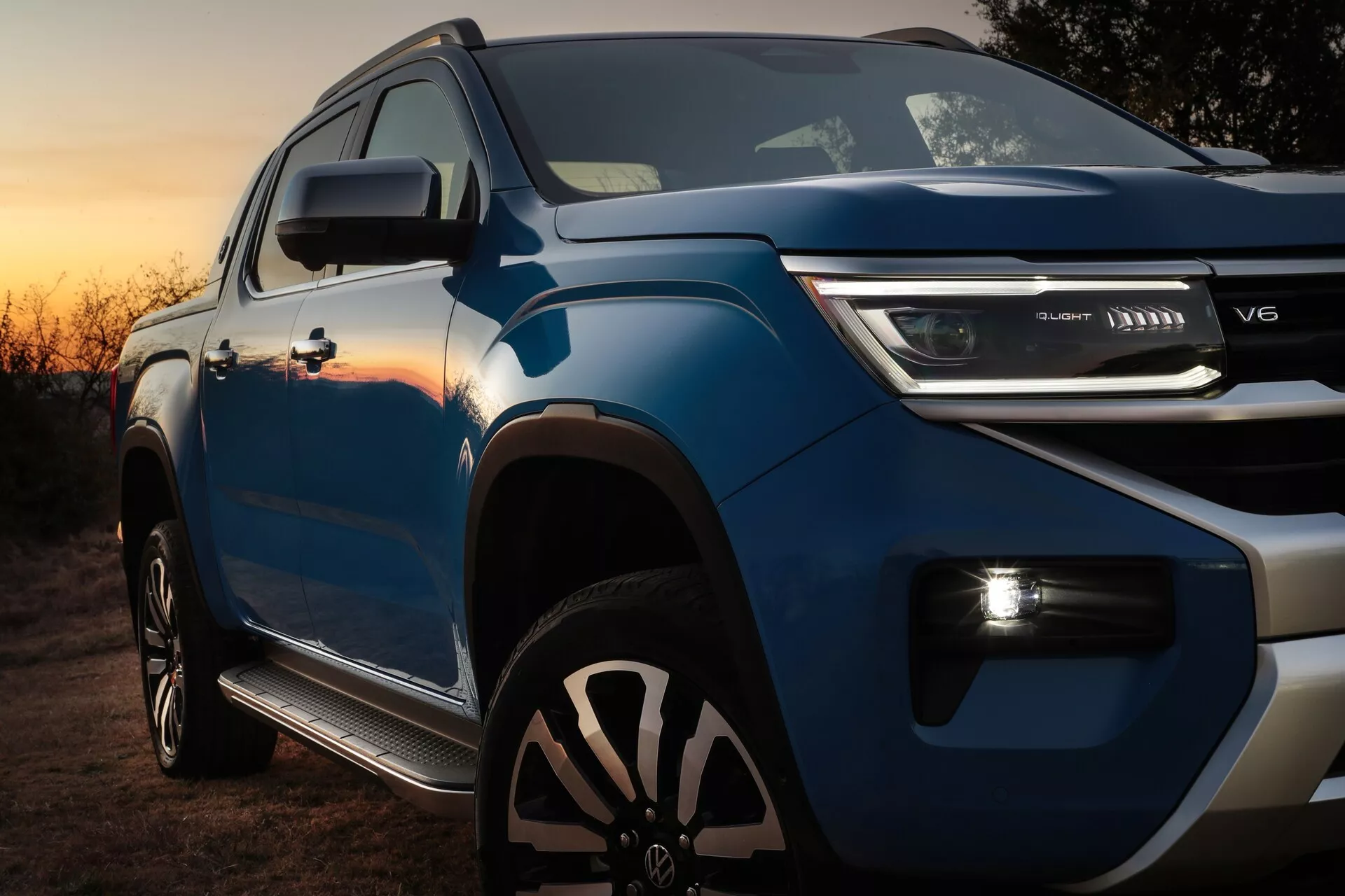 Vue rapprochée d'un pick-up bleu moderne, mettant en valeur sa hauteur et son design robuste de la partie avant, capturé sous un angle bas. La perspective du quart avant révèle une calandre contemporaine avec une disposition horizontale, un badge central et des feux de jour LED horizontaux élégants. Sous la calandre principale se trouve une importante prise d'air secondaire suggérant un moteur turbo. Le capot présente des lignes musclées surélevées et les rétroviseurs latéraux ont des indicateurs de signalisation intégrés. Le profil latéral visible inclut une bosse de l'aile et de grandes roues en alliage noir à rayons multiples. La photo, prise au lever ou au coucher du soleil, montre un pick-up garé à l'extérieur, mettant en avant son apparence robuste et puissante.