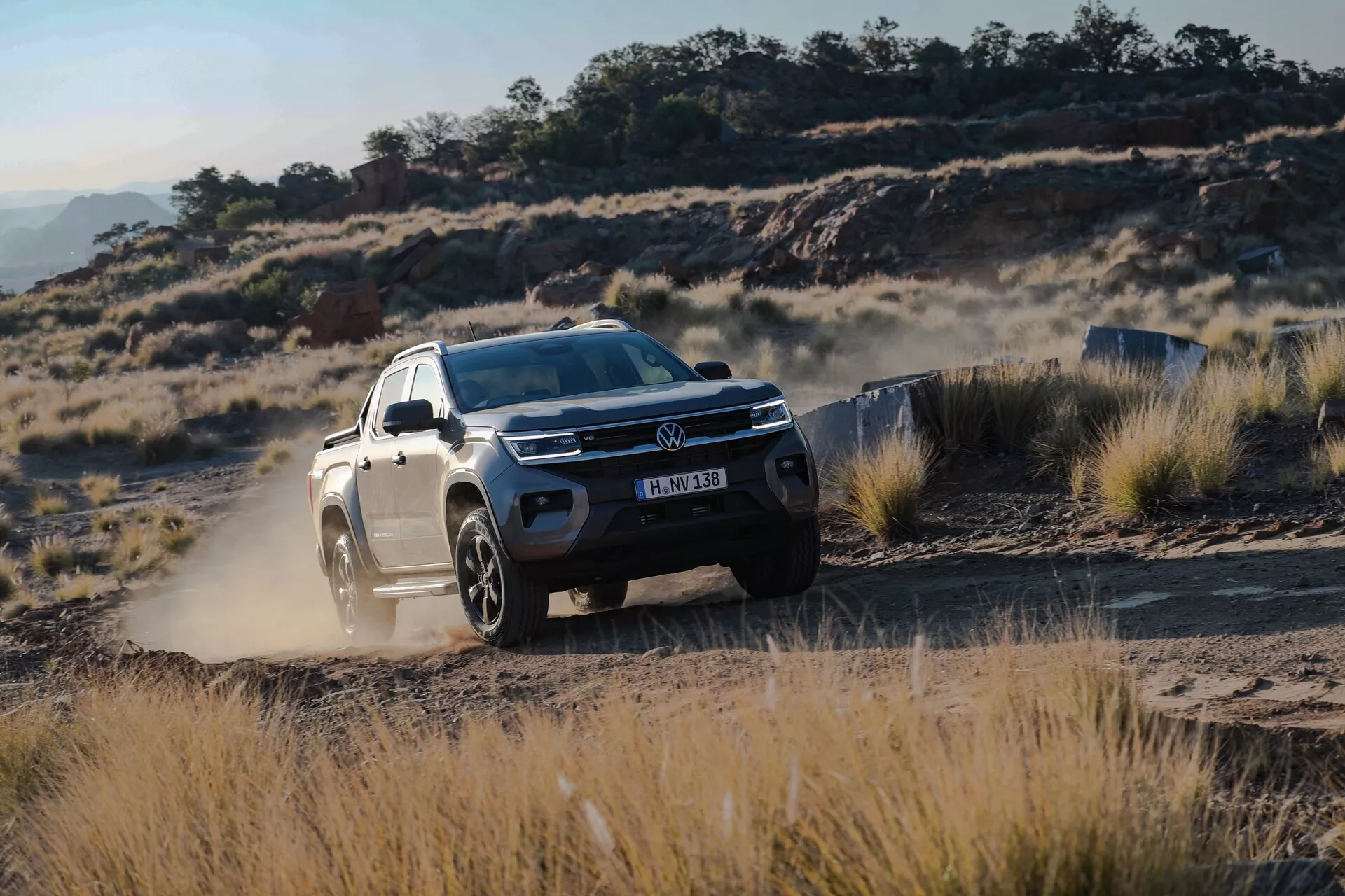 Prise de vue dynamique du SUV VW Nutzfahrzeuge Amarok 2023 de 2ème génération dans un environnement tout-terrain. L'image capture une vue de côté avant trois quarts, mettant en avant la calandre, les phares, le pare-chocs avant et le capot du véhicule, ainsi que le côté conducteur du pare-chocs avant à la porte arrière. Le SUV, peint dans une couleur claire, contraste avec le fond rugueux des sentiers poussiéreux, des herbes sèches et du terrain rocheux. La hauteur de caisse surélevée et les grandes roues en alliage soulignent ses capacités tout-terrain. Un nuage de poussière soulevé par les pneus arrière suggère un mouvement rapide. Les phares LED et les feux de jour sont clairement visibles, mettant en avant sa technologie d'éclairage moderne. L'insigne du fabricant, le design de la calandre et la texture de la poussière sont distinguables, accentuant le design élégant et l'apparence robuste du véhicule.
