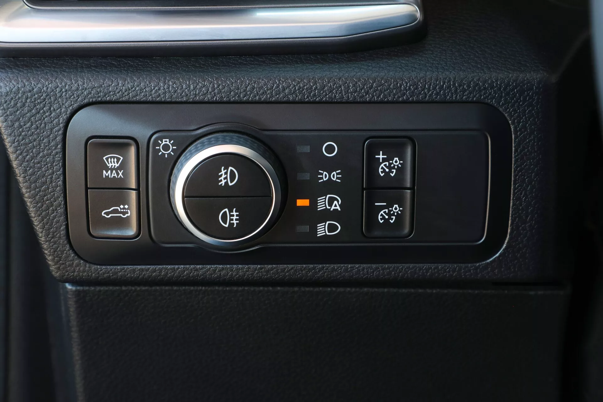 Vue rapprochée du panneau de commande des phares du VW Nutzfahrzeuge Amarok 2023 (2e génération). Ce panneau rectangulaire aux coins arrondis est intégré dans un tableau de bord texturé noir. Il comporte un bouton à gauche pour les phares et les feux de stationnement, y compris les phares complets, les feux de jour/de stationnement, le mode AUTO et Off. Les boutons adjacents contrôlent le dégivreur du pare-brise avant, le dégivreur de la lunette arrière, les rétroviseurs chauffants et le système auto-stop/start. Les symboles et lettres rétroéclairés, ainsi que les surfaces tactiles, assurent une utilisation facile et une bonne visibilité en basse luminosité.