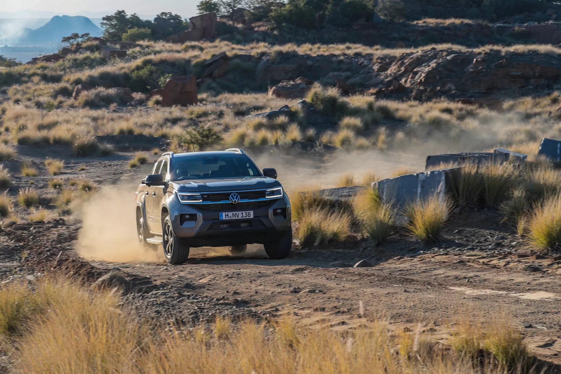SUV Volkswagen Amarok 2023 roulant sur un chemin de terre, vue de trois quarts avant montrant les phares LED, la grille détaillée avec l'emblème et le design des roues sur un arrière-plan tout-terrain accidenté avec une traînée de poussière.