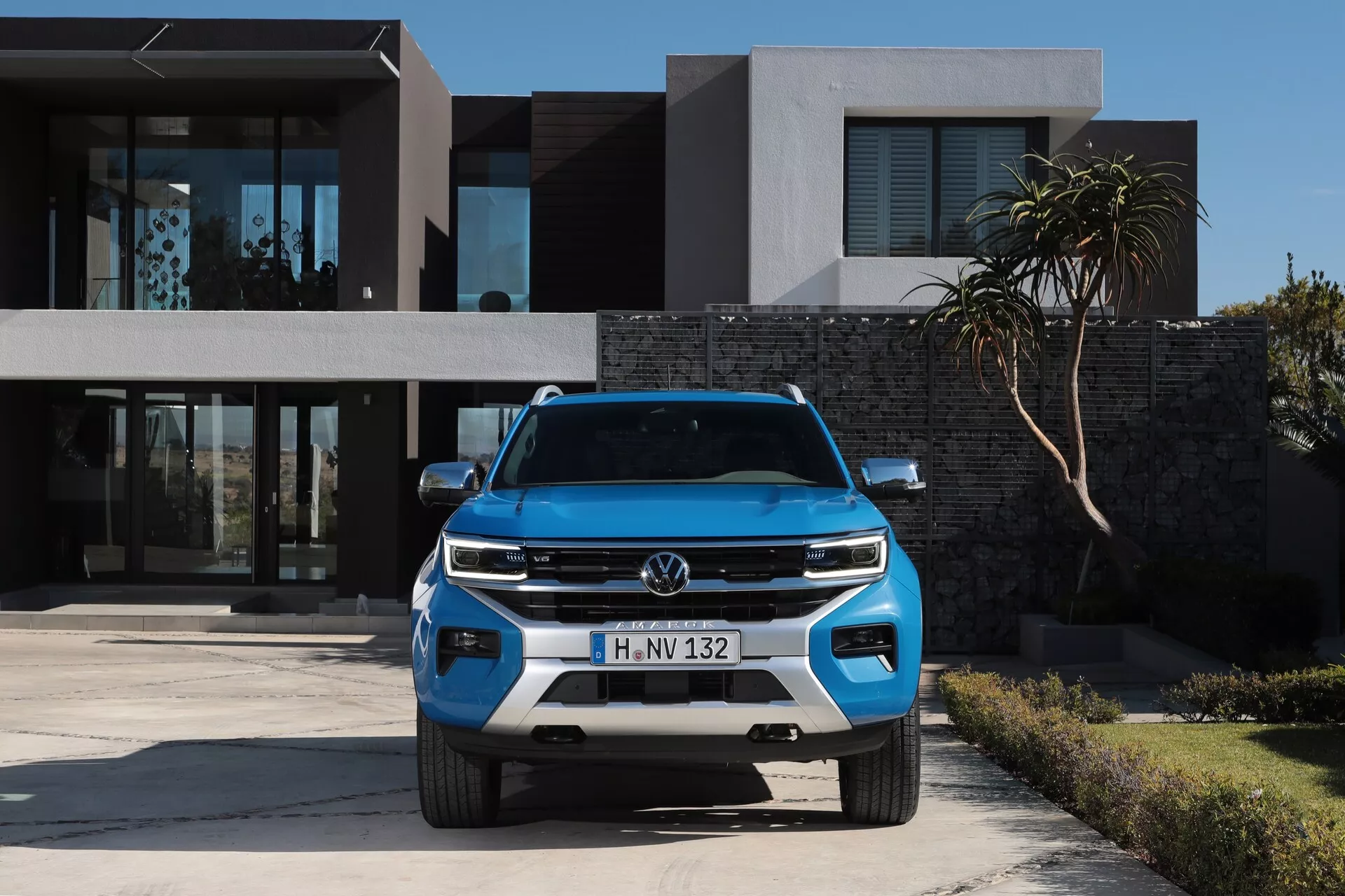 Vue de face d'un SUV Volkswagen Amarok 2023 bleu garé devant un bâtiment résidentiel moderne, mettant en valeur les phares élégants du véhicule, le design de la calandre, les feux de jour, le logo Volkswagen bien visible, les capteurs du pare-chocs inférieur et le capteur radar, soulignant ses caractéristiques de design, d'éclairage et de technologie de sécurité.