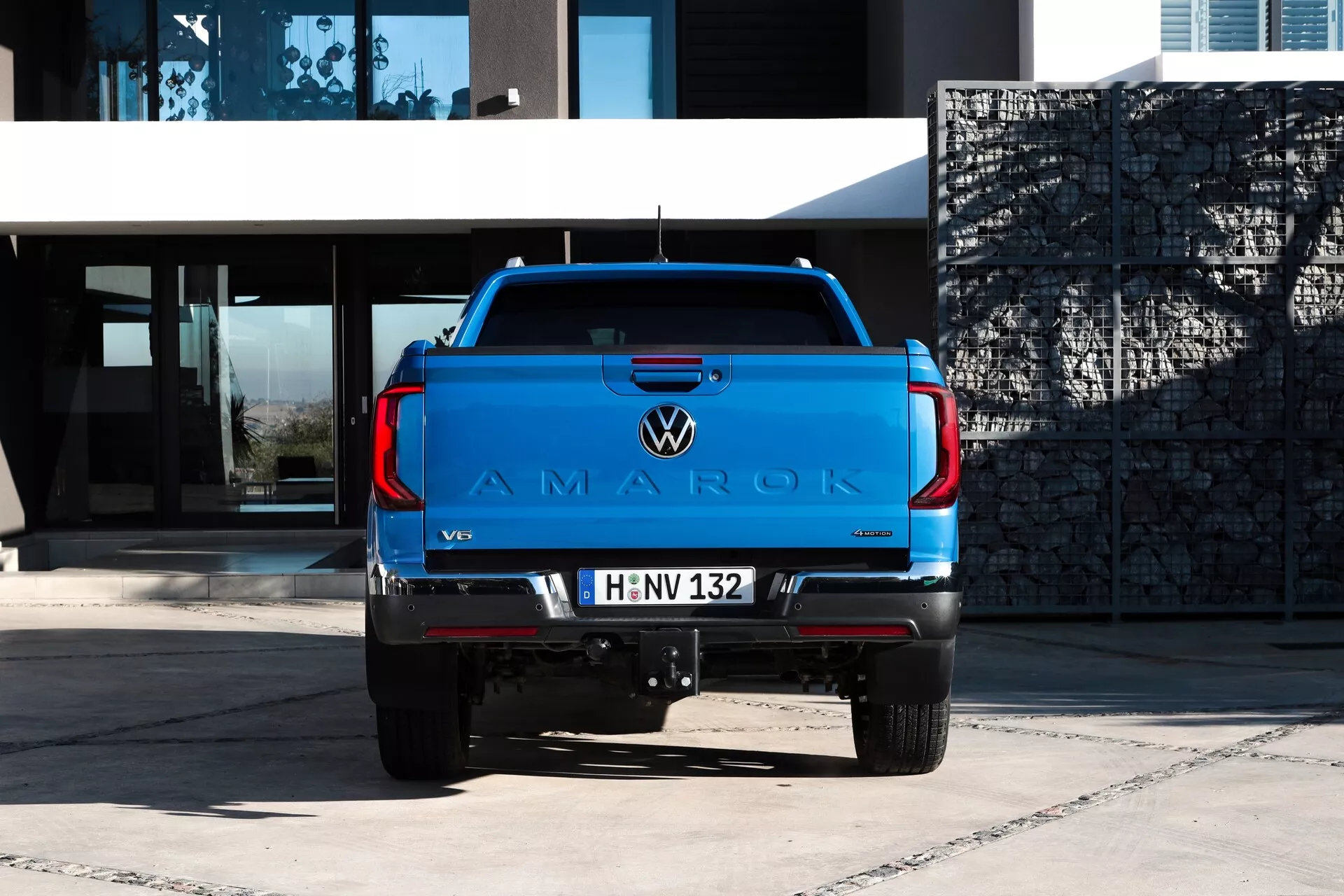 Vue arrière d'un pick-up Volkswagen Amarok 2023 avec une peinture bleue vibrante sur une surface pavée, montrant le grand logo 'AMAROK' sur le hayon, l'emblème VW, le badge moteur V6, les feux arrière modernes, l'attelage de remorque, les capteurs de stationnement, la plaque d'immatriculation de style européen et le système d'échappement double, sur un fond de bâtiment moderne à la lumière naturelle du jour.