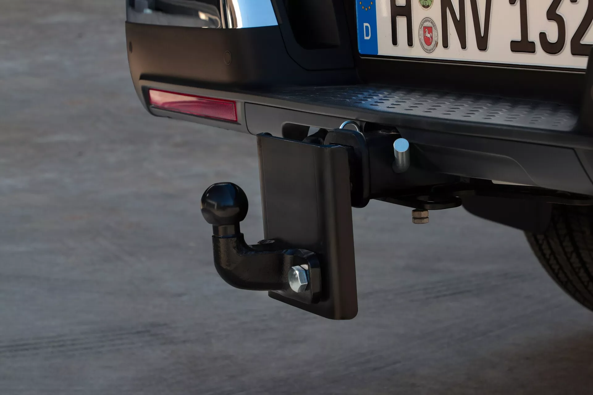 Vue rapprochée de l'attelage de remorquage du VW Nutzfahrzeuge Amarok 2023 de deuxième génération avec insistance sur la section découpée du pare-chocs arrière et le mécanisme de l'attelage, comprenant un récepteur carré avec une barre de remorquage, une goupille de sécurité et un boulon horizontal, plaque d'immatriculation arrière partiellement visible et feu antibrouillard arrière, stationné sur asphalte avec éclairage naturel.