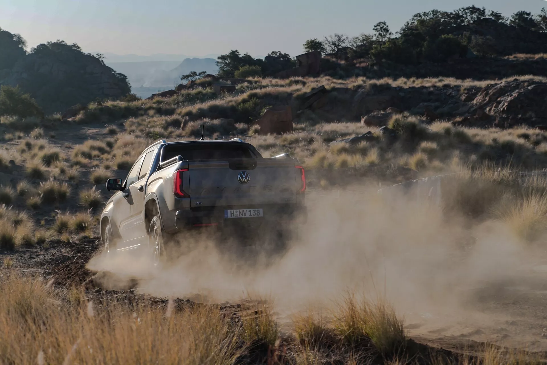 Moment dynamique tout-terrain du pick-up Volkswagen Amarok 2023 de deuxième génération roulant sur un sentier poussiéreux, avec un nuage de poussière enveloppant l'arrière. Photo prise sous un angle de trois quarts arrière, montrant la benne, la roue arrière, le hayon et la carrosserie côté passager. Le camion est légèrement incliné, indiquant un terrain accidenté ou un virage rapide, mettant en avant ses capacités de transmission intégrale, de garde au sol et de suspension. L'éclairage chaud suggère le début de la matinée ou la fin de l'après-midi, positionnant le camion comme capable de gérer des conditions difficiles.