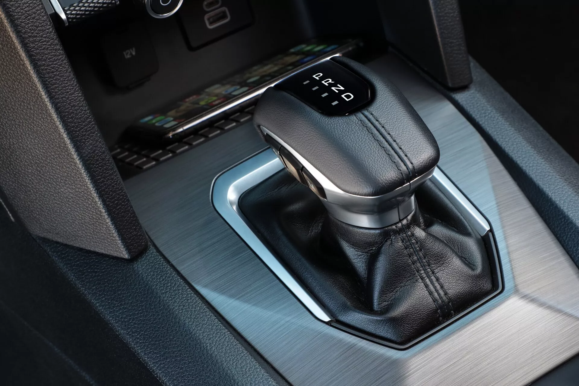 Gros plan du levier de vitesse automatique du VW Nutzfahrzeuge Amarok 2023, avec un pommeau en cuir et un indicateur de vitesse illuminé affichant 'P' pour Parking. L'angle de vue depuis le dessus et l'avant-droit révèle un cadre métallique avec un éclairage d'ambiance bleu et un petit compartiment de rangement dans la console centrale.