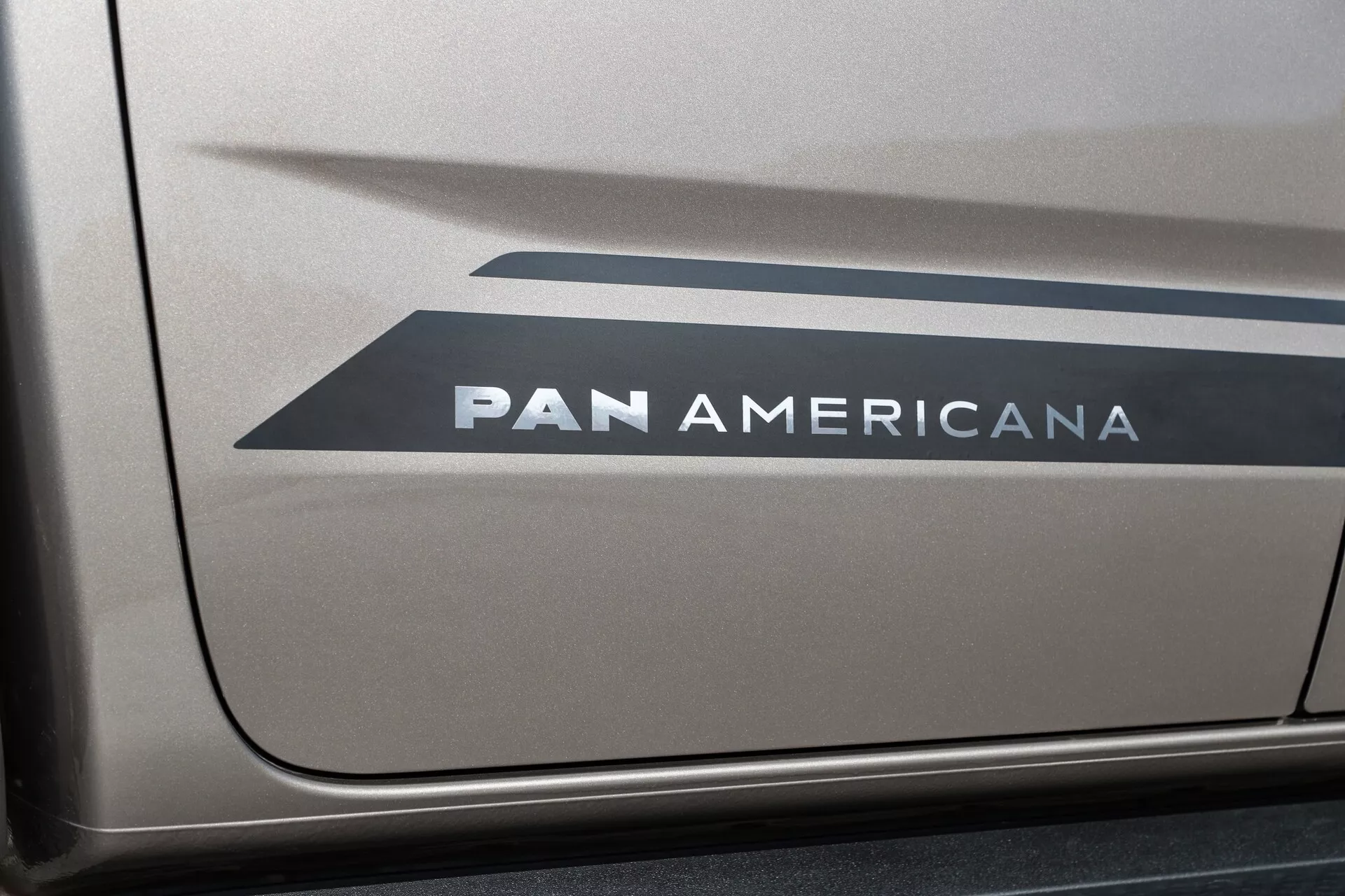 Vue latérale rapprochée d'une section du VW Nutzfahrzeuge Amarok 2023 (deuxième génération) montrant un graphique avec deux formes en chevron pointant vers l'arrière et le texte en gras 'PAN AMERICANA' sur une probable finition de peinture métallique, avec un éclairage clair et uniforme.