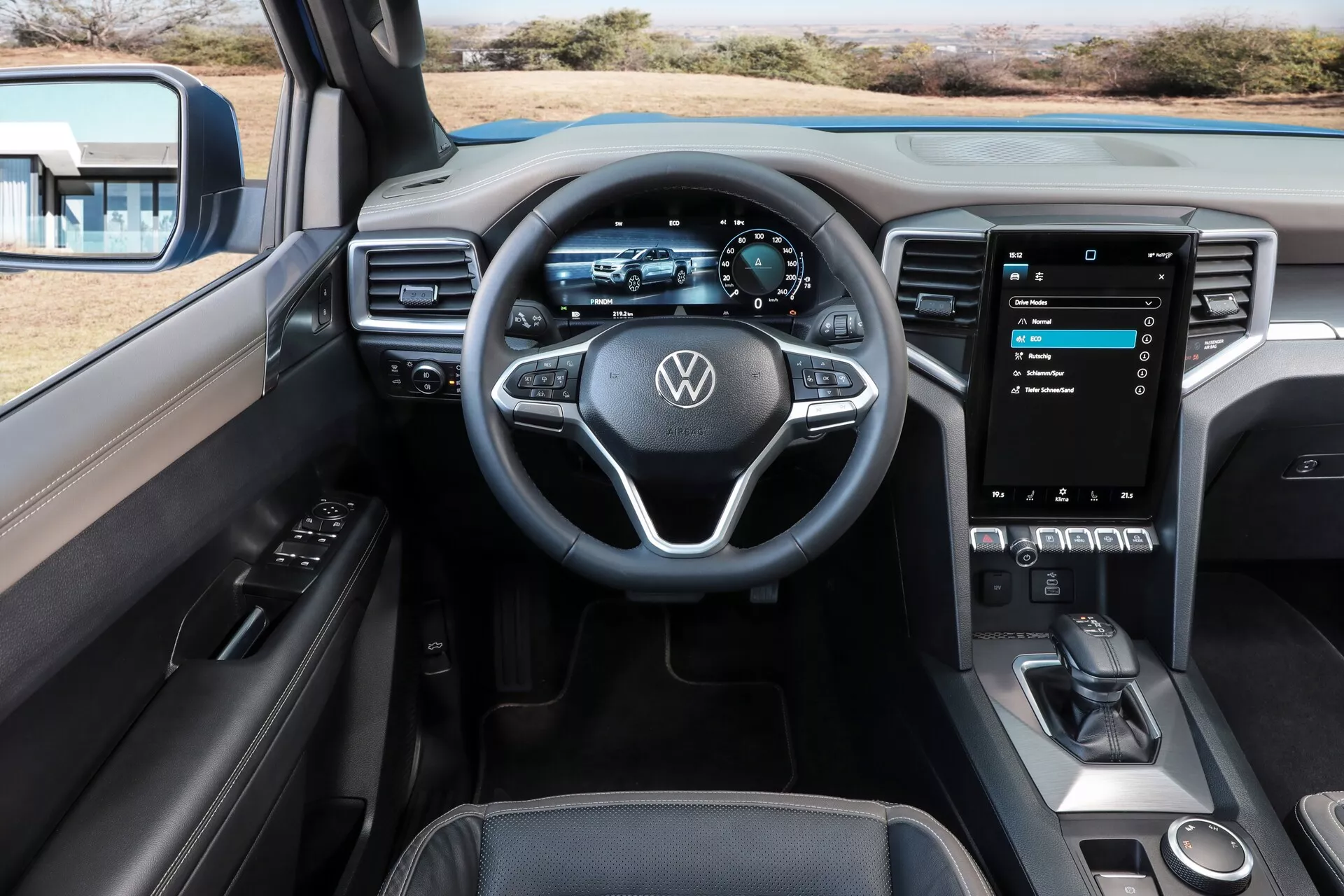 Vue intérieure de la Volkswagen Amarok 2023 (2e génération), montrant le cockpit du conducteur depuis la perspective du siège arrière. L'image met en avant le volant moderne à trois branches avec boutons multifonctions, le tableau de bord entièrement numérique, le grand système d'infotainment à écran tactile vertical affichant le menu principal, des commandes de climatisation minimalistes et un sélecteur de vitesse élégant dans la console centrale. Le panneau de porte propose des commandes pour les vitres électriques et les rétroviseurs extérieurs, renforçant l'attrait technologique et esthétique de l'intérieur du véhicule.