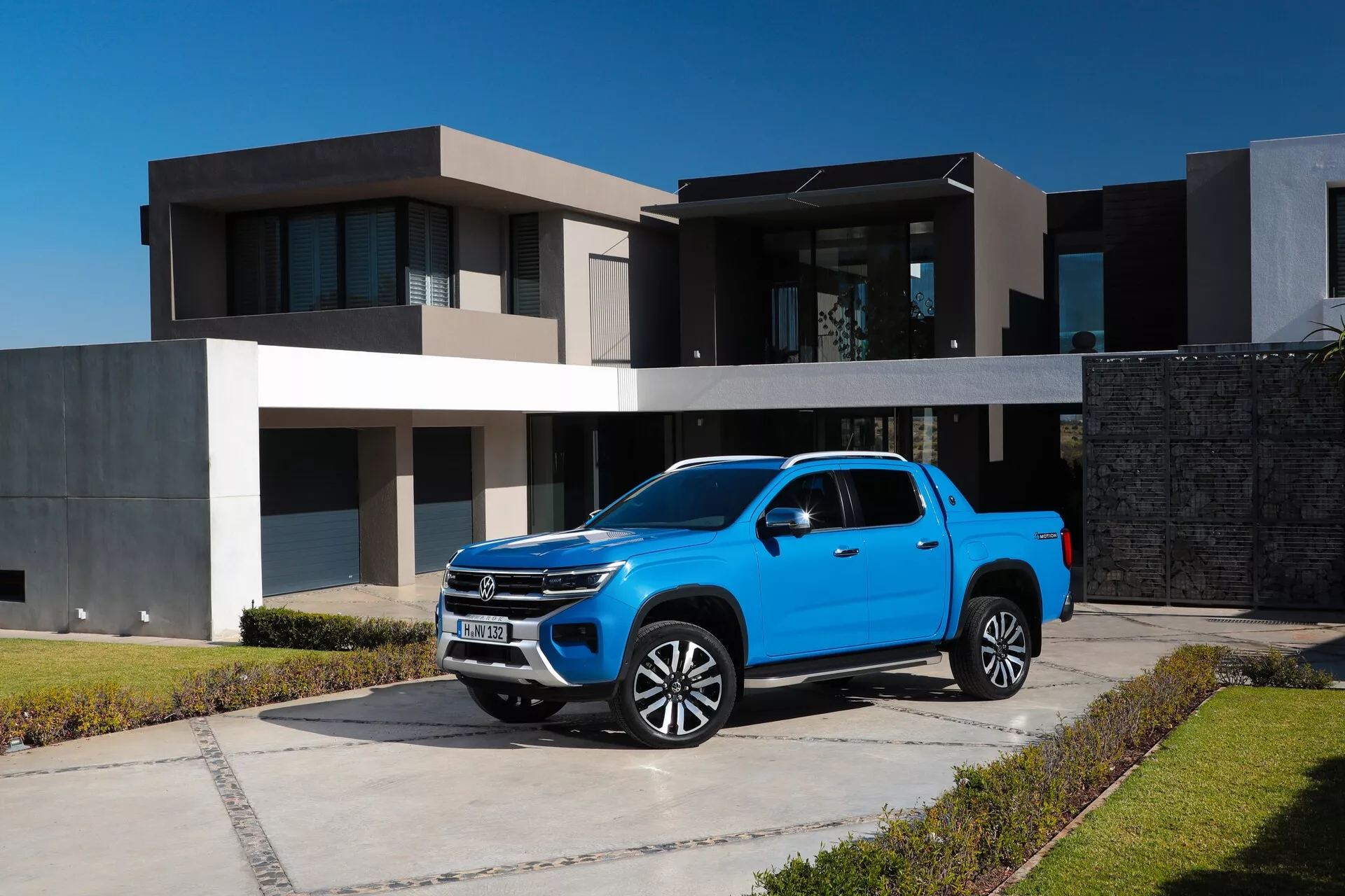 Pickup de deuxième génération VW Nutzfahrzeuge Amarok 2023 bleu vif garé sur une allée en béton devant une maison moderne, présenté sous un angle avant trois-quarts. Cette vue met en évidence la grande calandre avec une finition chromée, les élégants phares à LED, les jantes en alliage multi-rayons, la peinture brillante, le style de carrosserie double cabine, les rétroviseurs avec clignotants intégrés et les vitres légèrement teintées. La grande garde au sol et l'environnement suburbain sont également clairement visibles.