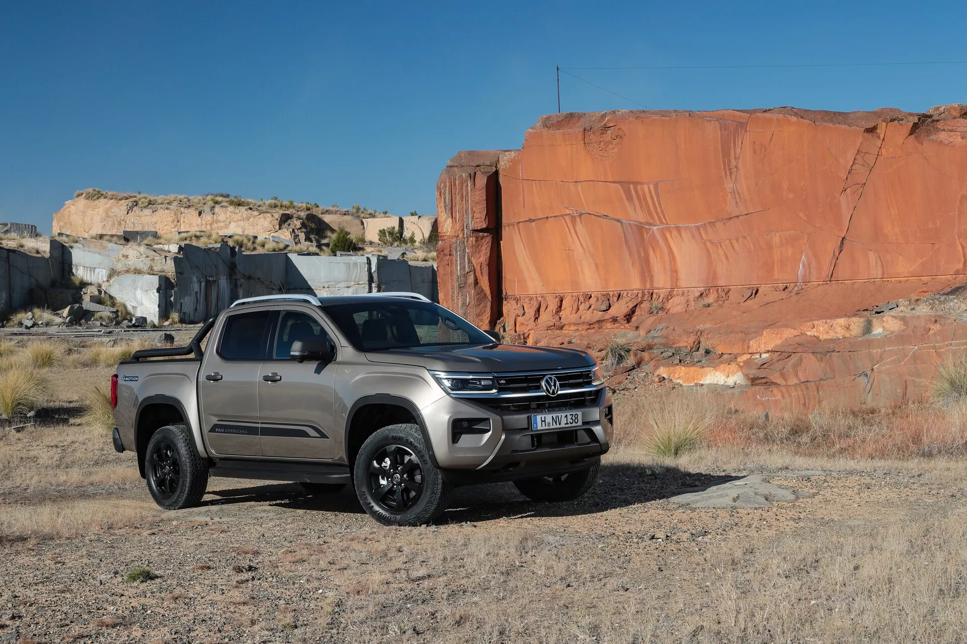 Un pick-up VW-Nutzfahrzeuge Amarok de deuxième génération de 2023 exposé dans un terrain rocheux naturel, mettant en avant ses capacités tout-terrain. La prise de vue en angle trois-quarts avant montre le pare-chocs avant de la couleur de la carrosserie avec des phares antibrouillard intégrés, un badge central proéminent sur la calandre, des unités de phares rectangulaires élégantes, des sections surélevées du capot, des roues en alliage noir et une garde au sol élevée. La configuration double cabine a quatre portes avec rétroviseurs latéraux, poignées de porte et marchepieds visibles. Le camion, positionné sur une surface plate en terre avec en toile de fond de grandes formations rocheuses rouges, est l'objet principal de la photographie prise en lumière naturelle.