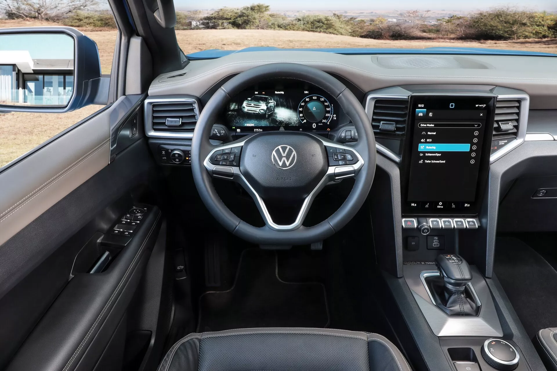 Vue intérieure depuis le siège du conducteur d'une Volkswagen Amarok 2023 (2ème génération), mettant en évidence le volant avec emblème Volkswagen et commandes multifonctions, tableau de bord entièrement numérique affichant les métriques du véhicule, système d'infodivertissement à écran vertical tactile, commandes HVAC à toucher sensible, levier de vitesse et esthétique luxueuse de cabine combinant cuir et accents métalliques.