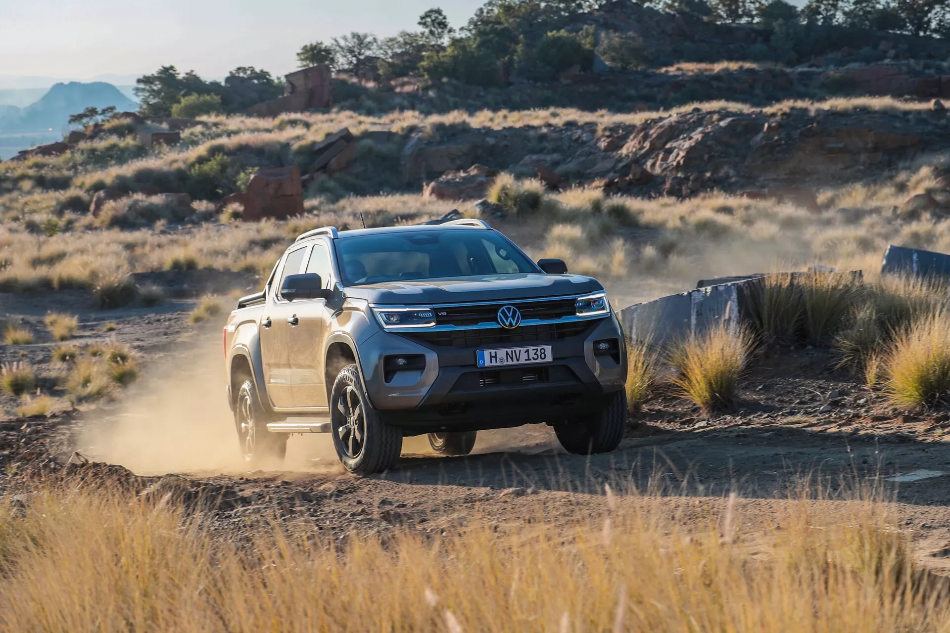 Vue de trois quarts avant du Volkswagen Amarok 2023 (2e génération). L'image met en évidence la calandre avant avec le badge Volkswagen, les phares élégants à LED, le pare-chocs avant avec prises d'air et partiellement le profil latéral côté conducteur, y compris la porte avant, l'aile, les arches de roue et les rétroviseurs latéraux. Le SUV moderne, avec calandre à lamelles horizontales, bande lumineuse à LED et roues en alliage tout-terrain, est capturé en mouvement sur un terrain rocheux et herbeux, montrant ses capacités tout-terrain et ses fonctionnalités avancées d'assistance à la conduite comme le régulateur de vitesse adaptatif et le freinage d'urgence autonome.