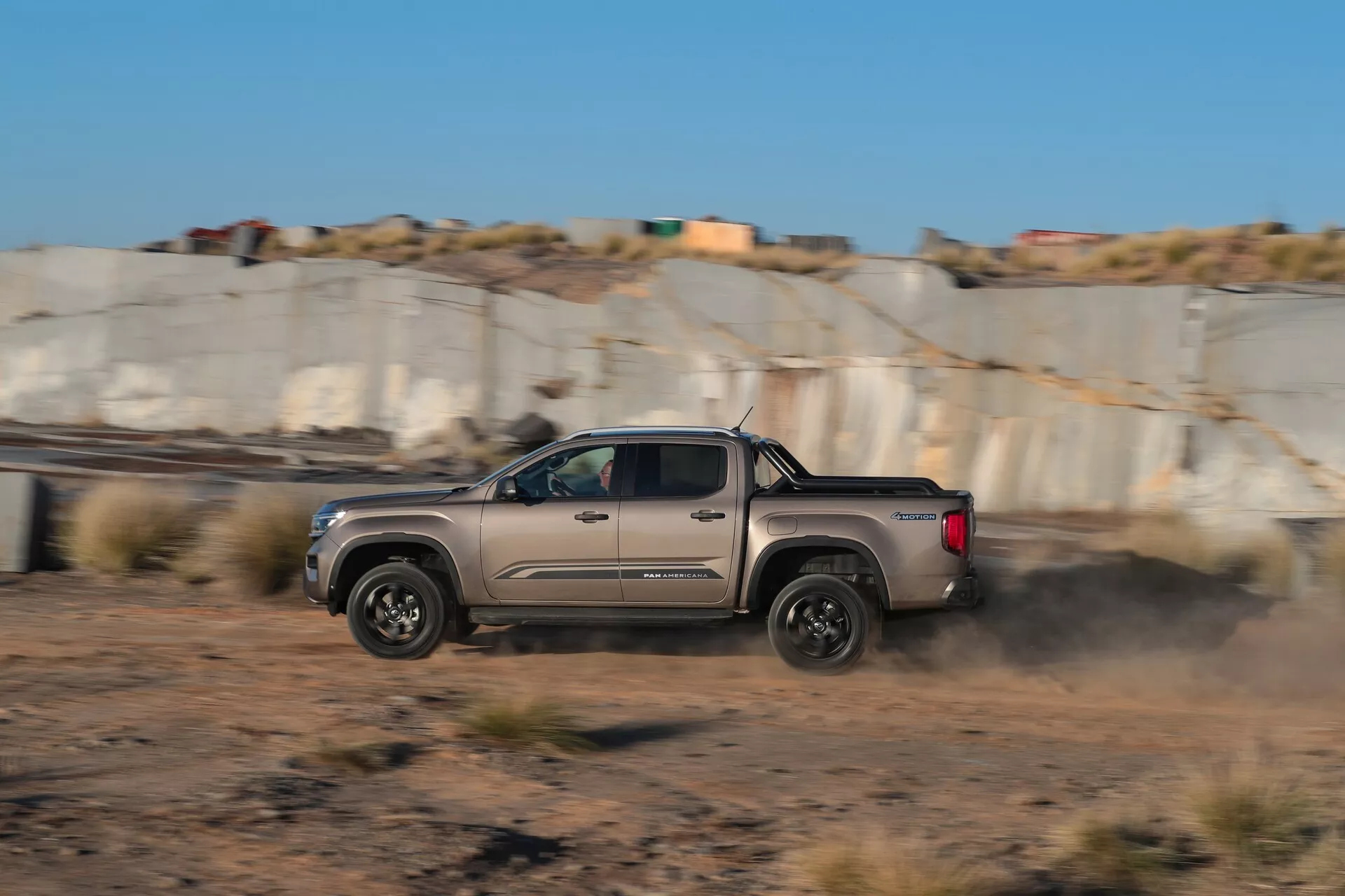 Vue de profil latéral du pick-up Volkswagen VW Nutzfahrzeuge Amarok 2ème génération 2023, capturé en mouvement avec un arrière-plan flou et un nuage de poussière derrière, soulignant sa manœuvre dynamique sur une route de terre. Doté d'une cabine double, de jantes en alliage et d'un design moderne et aérodynamique, indicatif de capacités tout-terrain avancées.